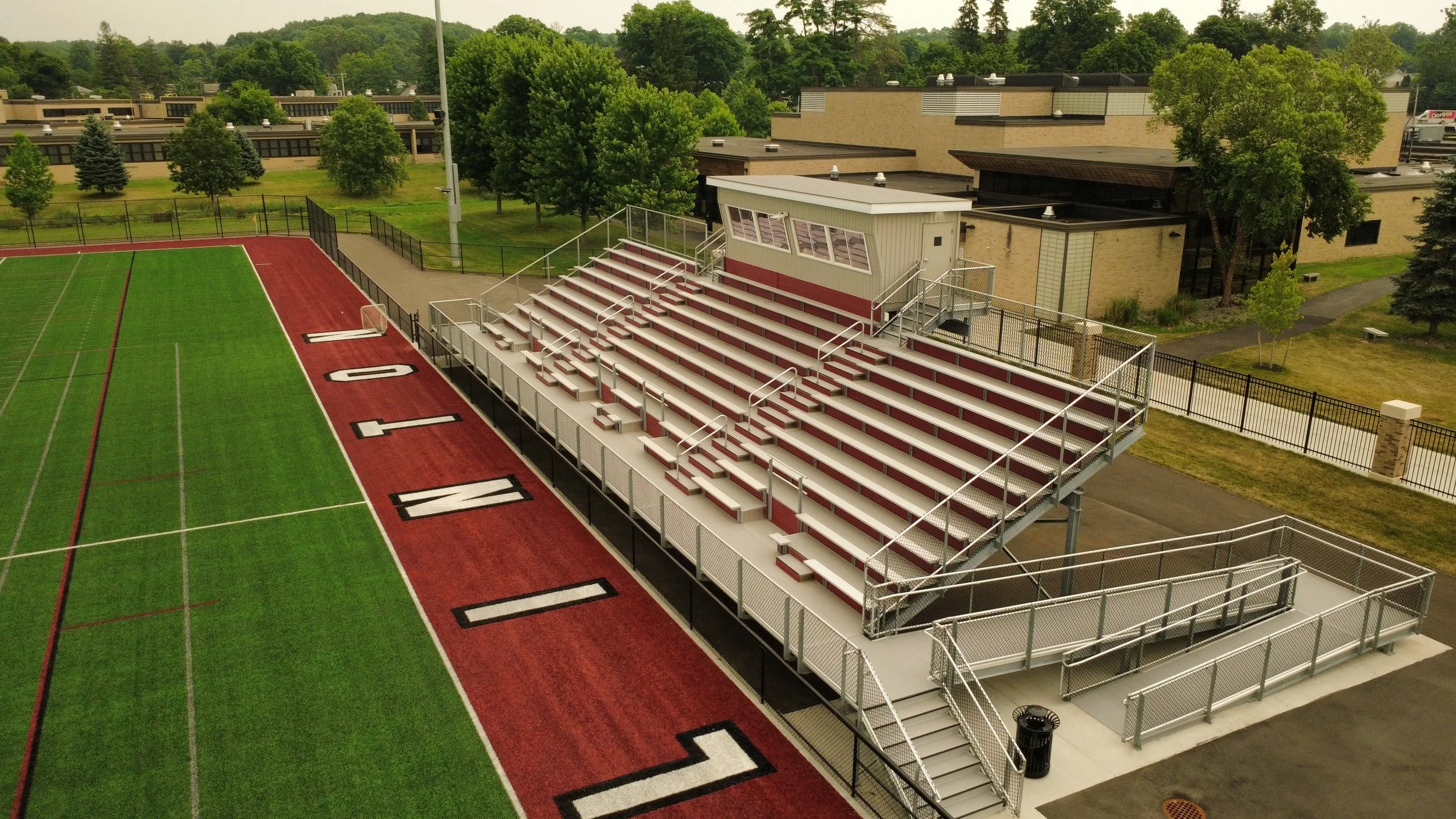 View of a school football field with red running track, bleachers, and trees in the background.