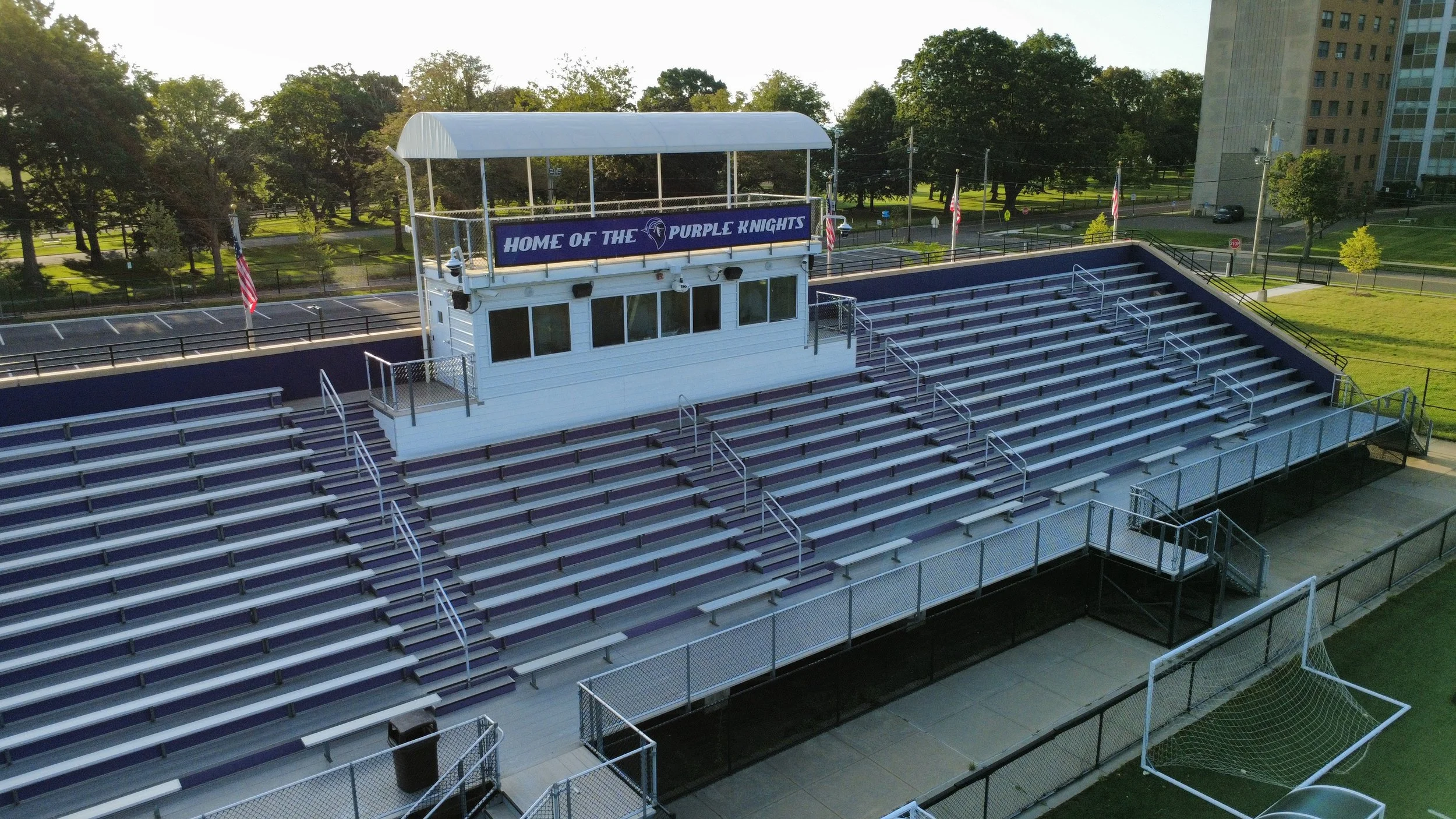 Empty purple and white bleachers at a sports stadium with a white press box that has a sign reading 'Home of the Purple Knights' and American flags. Green trees and tall buildings are visible in the background.