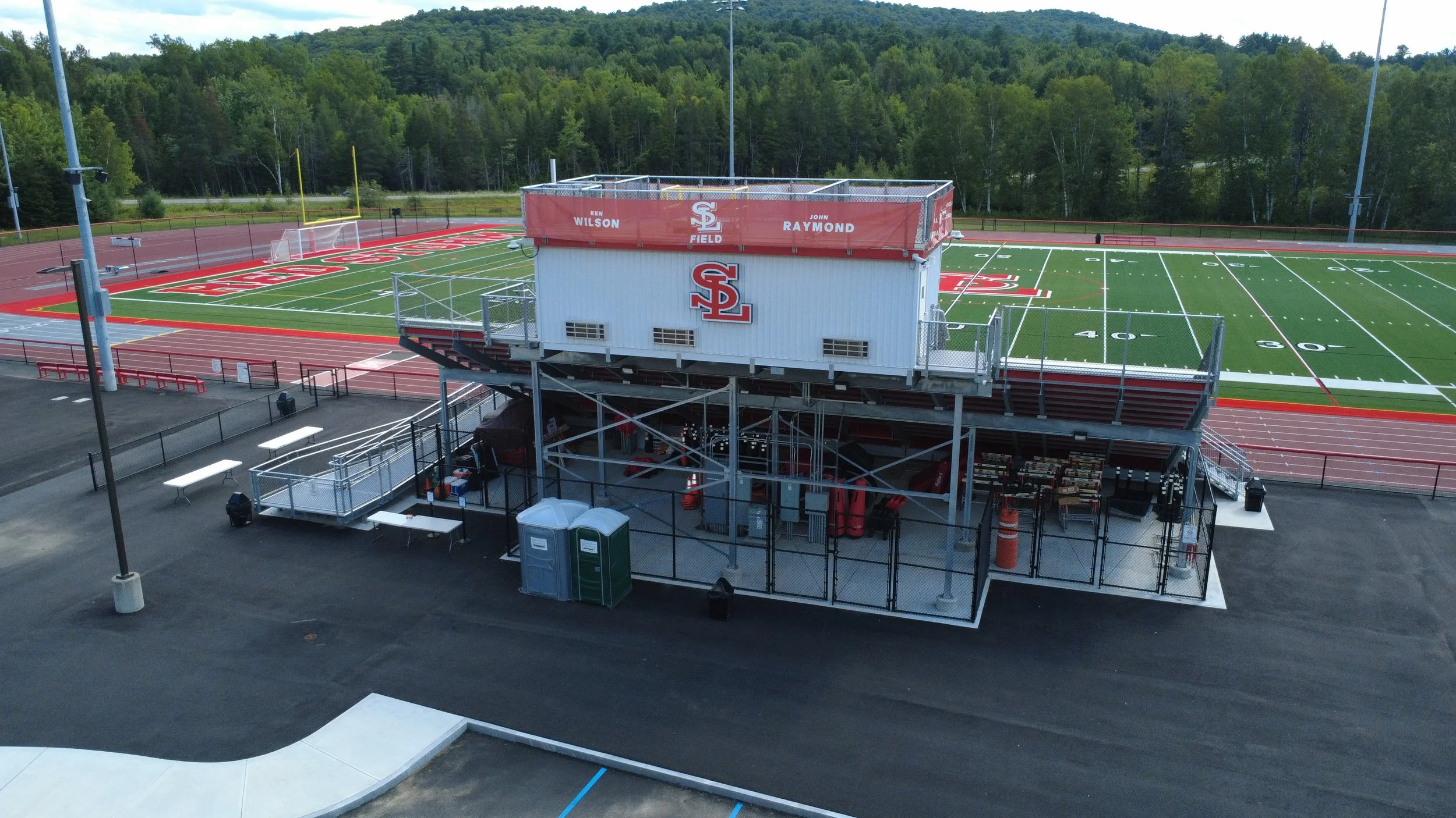 A football field with artificial turf, surrounded by a running track, and an elevated white press box with red accents in the center. Tall trees in the background.
