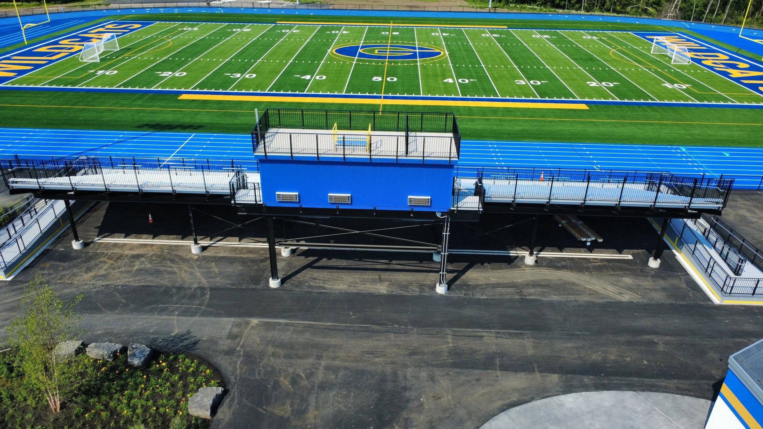 An empty football field with blue and yellow markings, viewed from above, with a track surrounding it and a blue press box structure in the foreground.