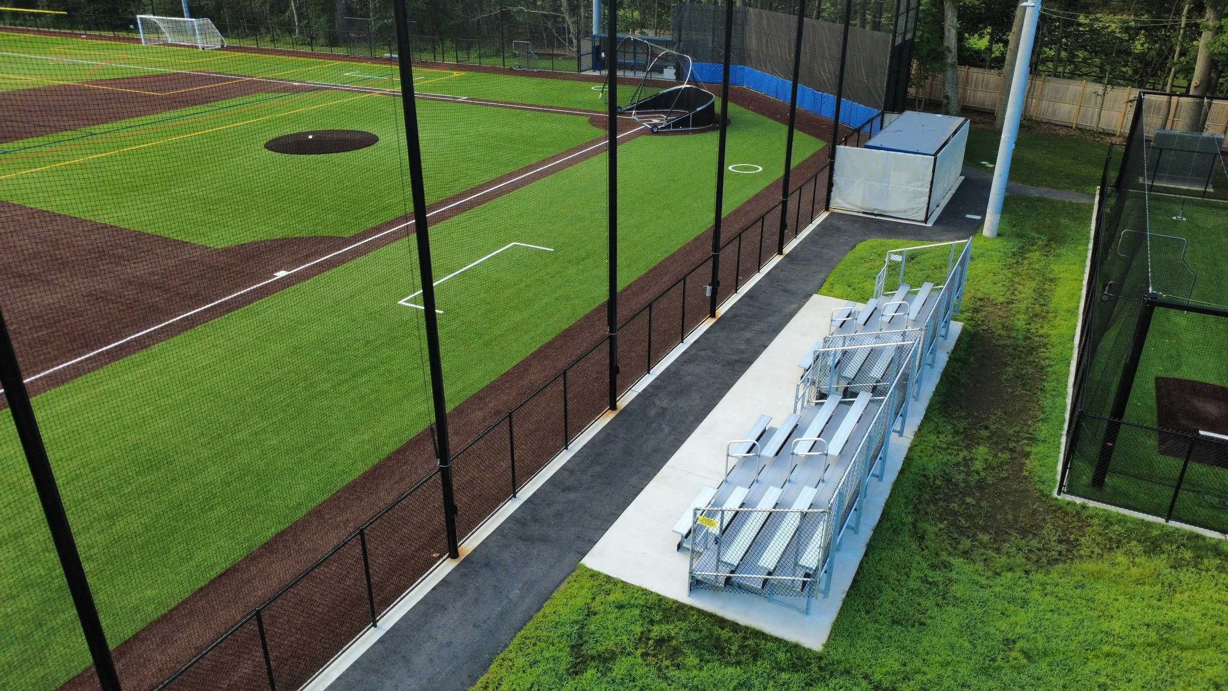 A baseball field with a dirt infield, green outfield grass, and a safety net surrounding it, seen from an elevated perspective. There are bleachers and a small covered area for spectators, and a few trees and a fence in the background.