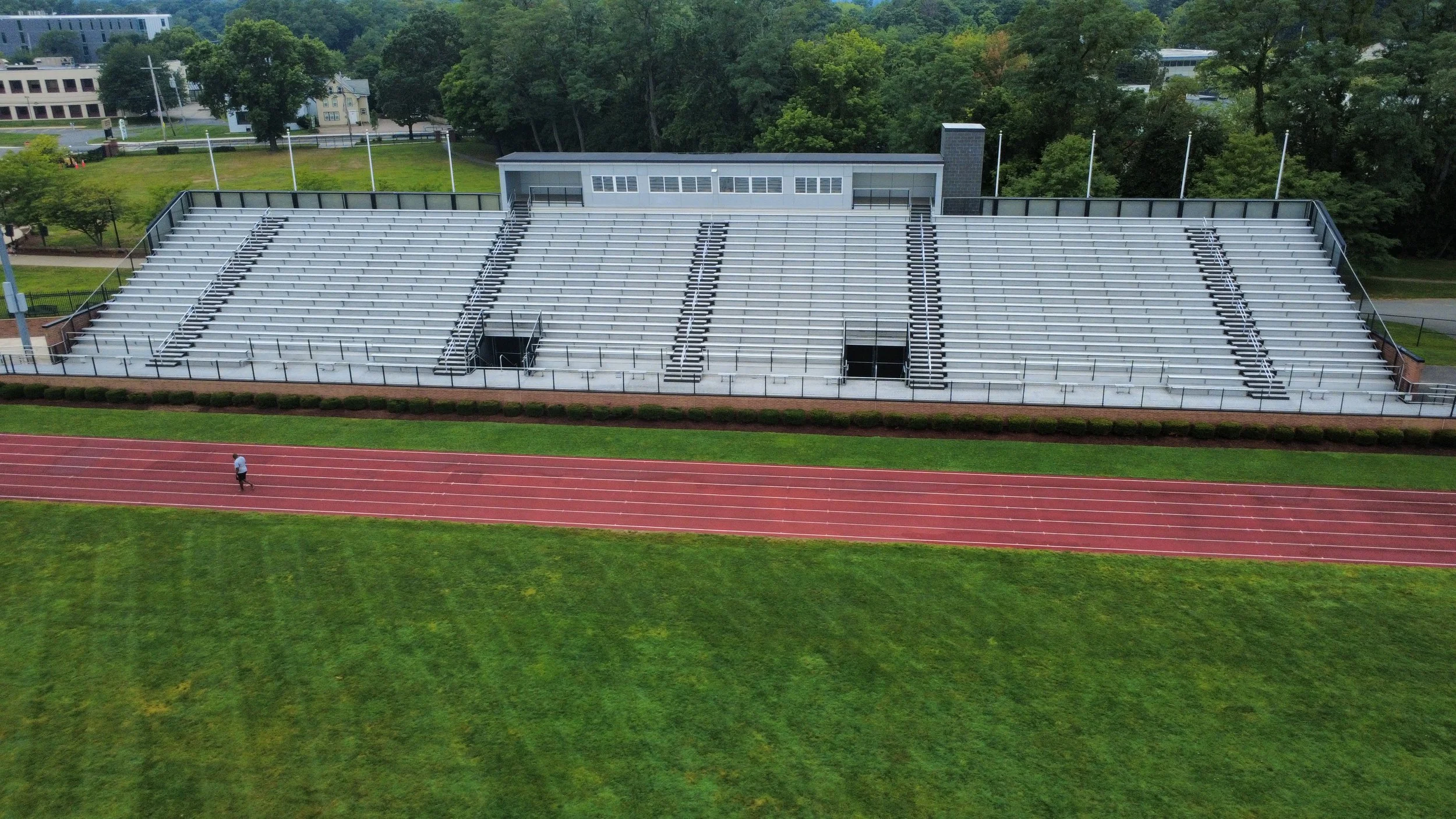 Aerial view of a sports stadium with empty bleachers and a red running track surrounding a green field. There is a single person walking on the track.