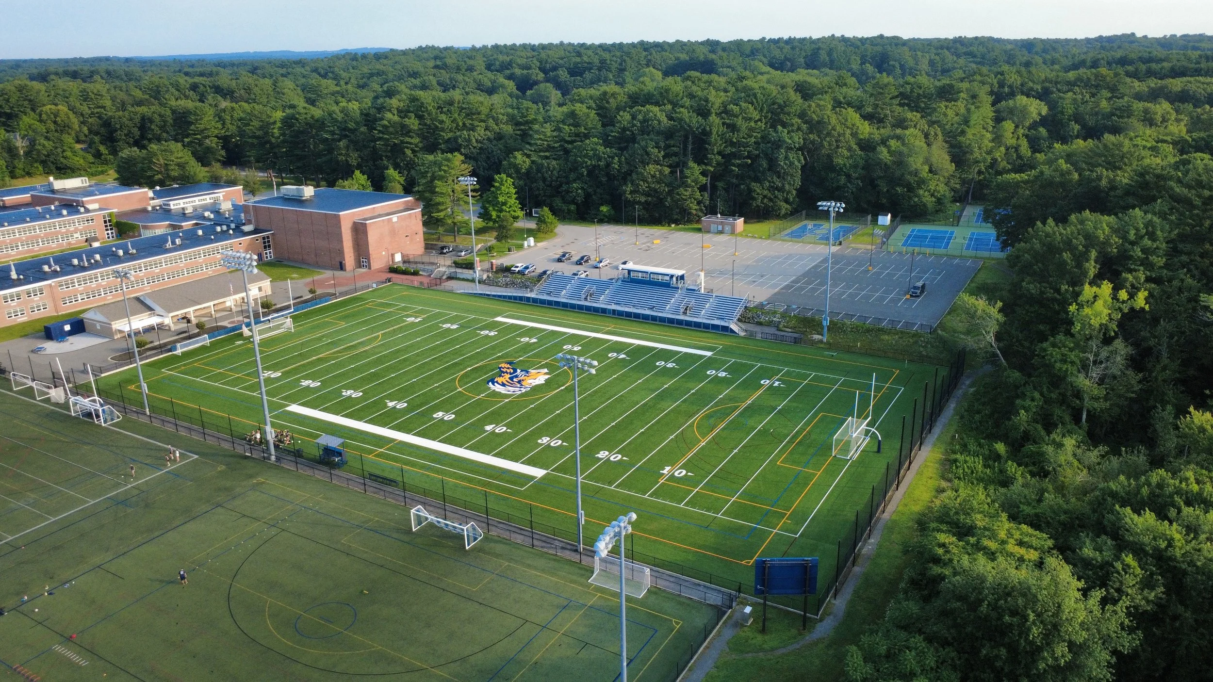 An aerial view of a school campus showing a football field with a tiger mascot logo, multiple tennis courts, parking lot, and school buildings surrounded by trees.