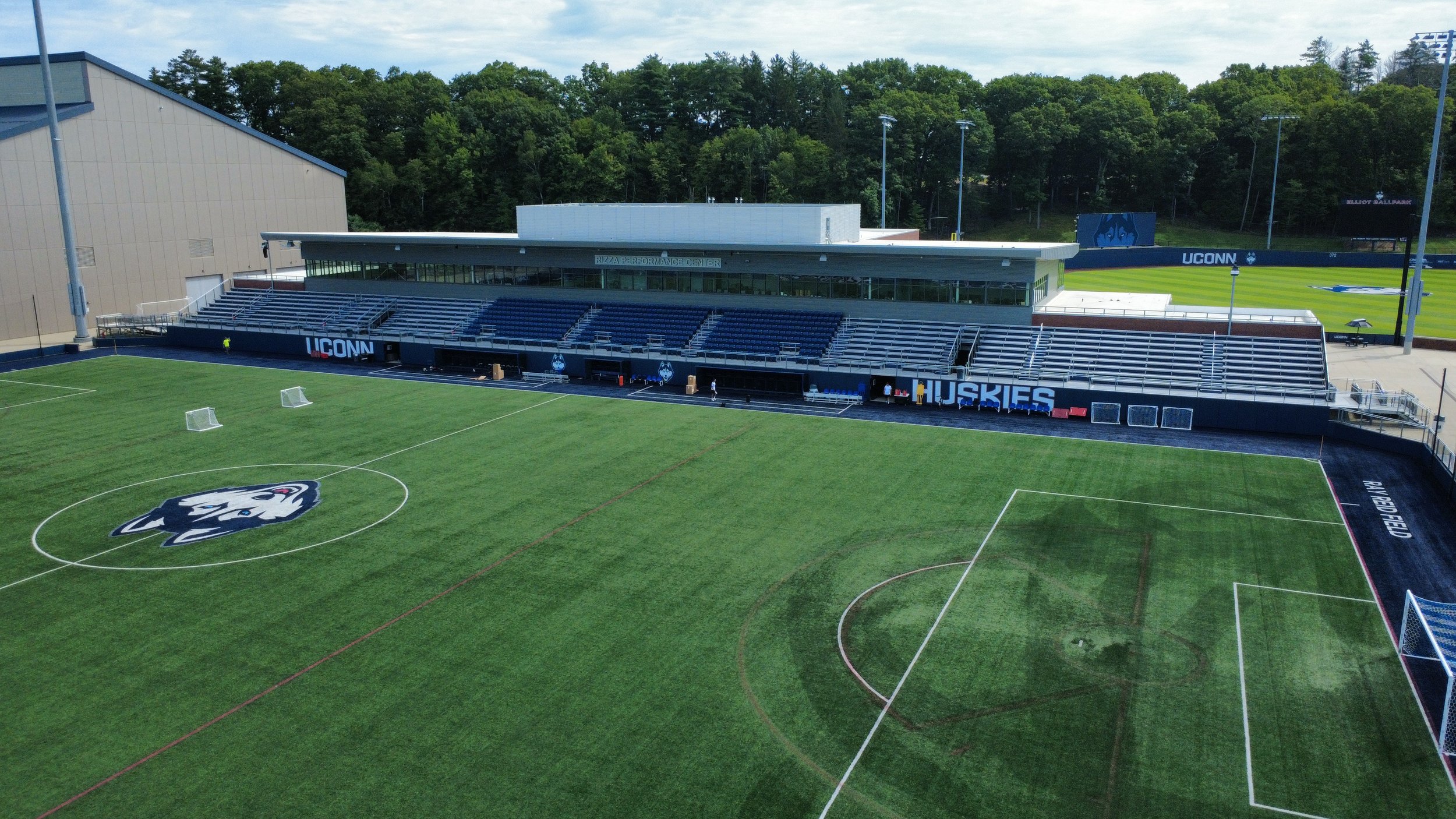 An empty soccer stadium with a field that has the University of Connecticut Huskies logo at the center, sideline seating area adorned with the Huskies name, and a press box labeled 'Rizza Performance Center,' surrounded by trees and light poles.