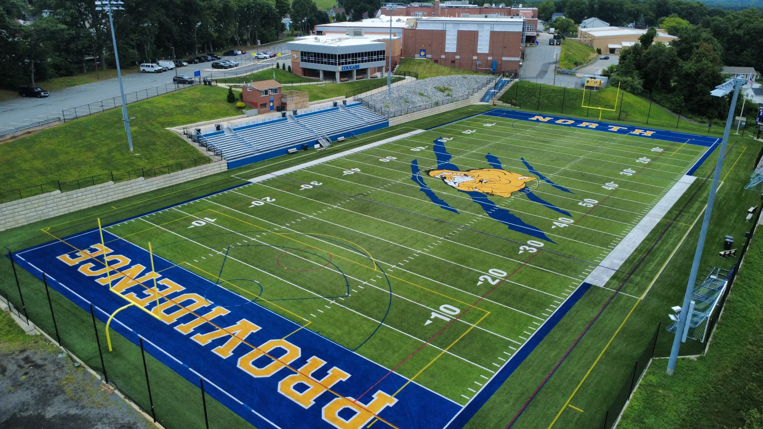 A football field with markings, a large logo of a bulldog in the center, and blue end zones reading 'NORTH' at one end and 'SOUTH' at the other. Surrounding the field are bleachers, a small building, and a parking lot with cars. Trees and school buil
