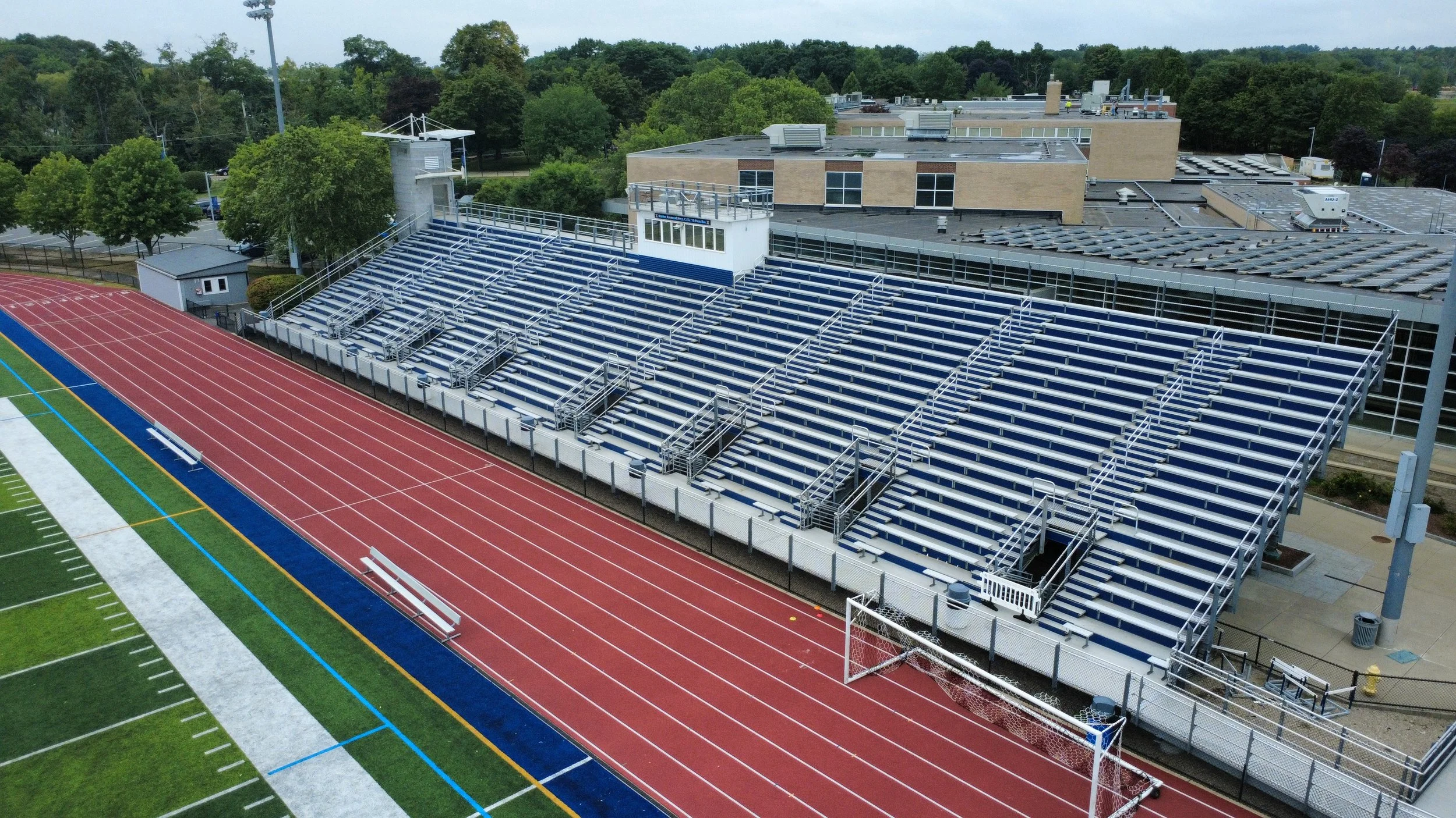 Aerial view of a running track and bleachers at a school or stadium with trees and buildings in the background.
