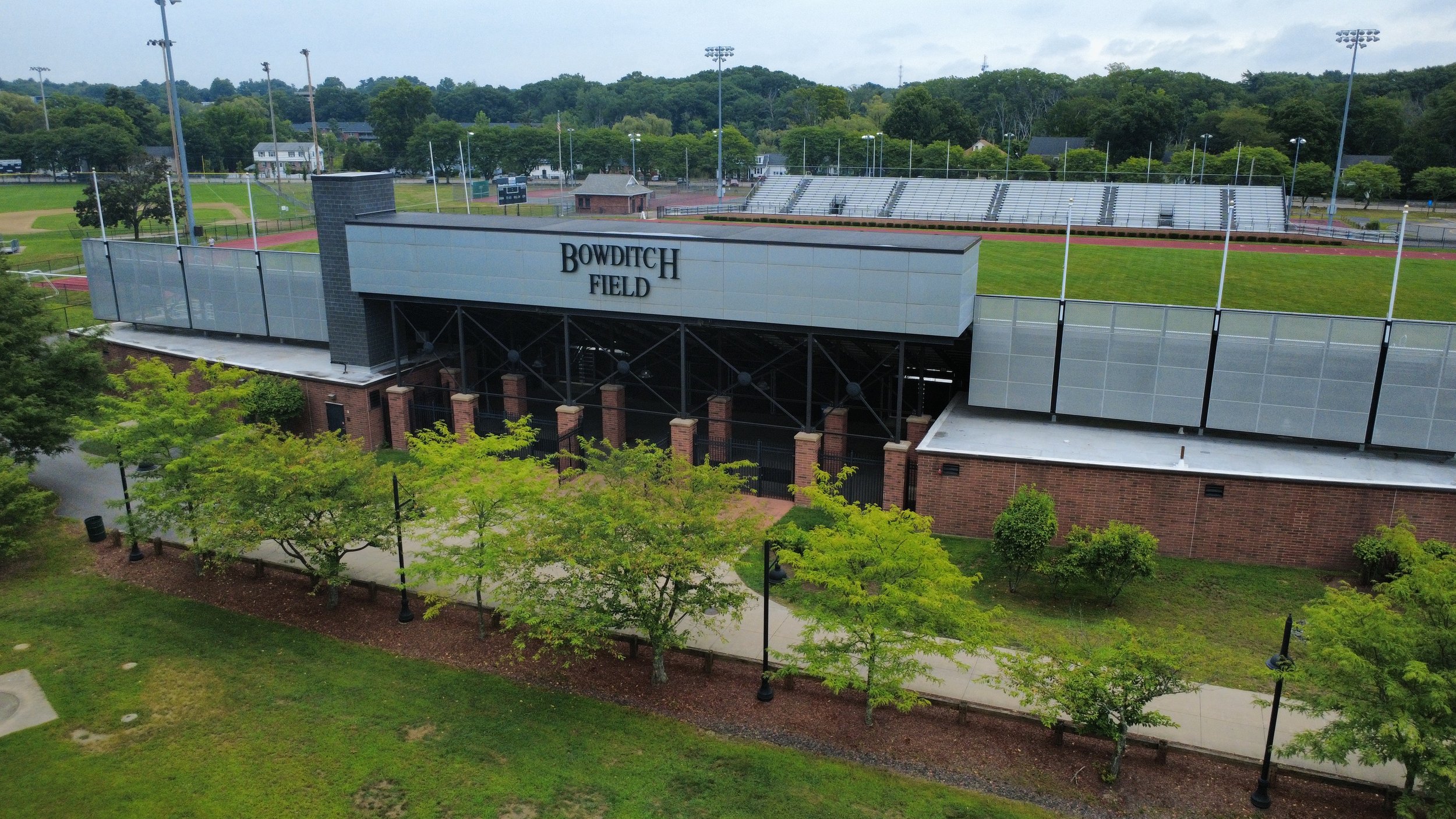 An outdoor sports field with a running track, bleachers, and a building labeled 'BOWDICH FIELD' in the foreground. Surrounding trees and a hill in the background.