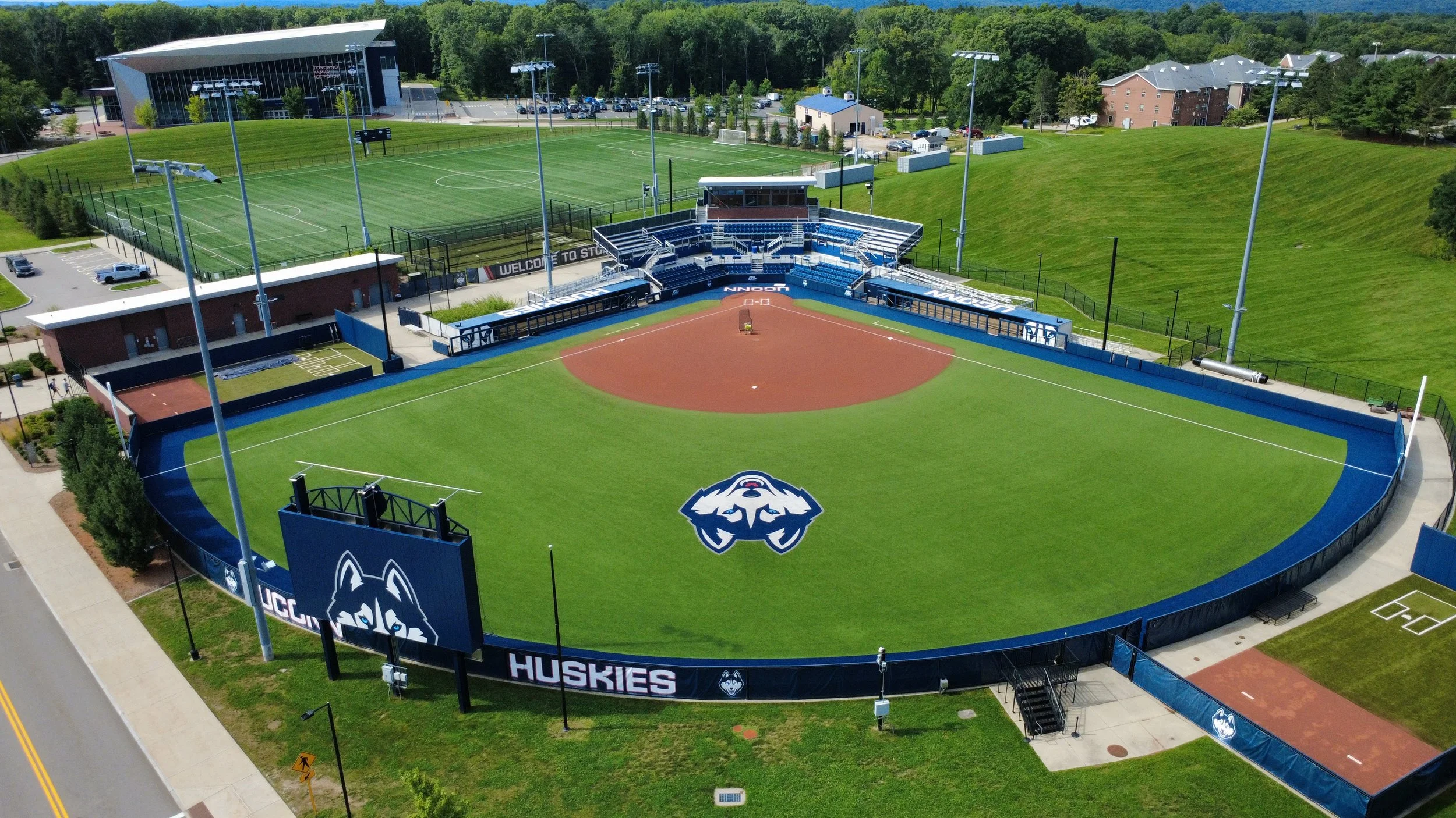 Aerial view of a baseball stadium with a green field and seating area, surrounded by a fence and lit by tall floodlights. Adjacent to the stadium is a soccer field with bright green grass and a large blue and white Husky logo at home plate. There are