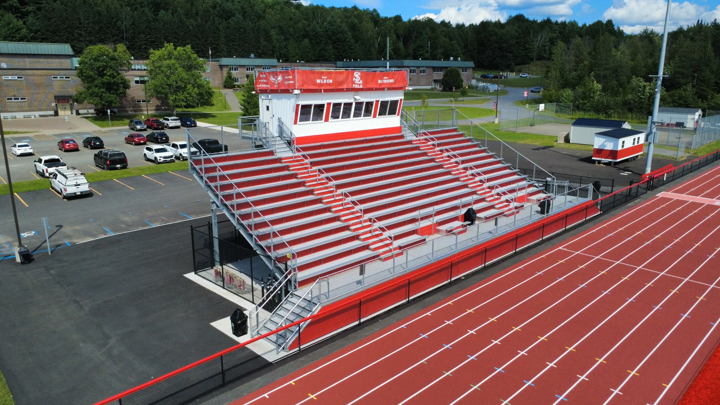 Red and white bleachers at a sports field with a small building and parking lot nearby.