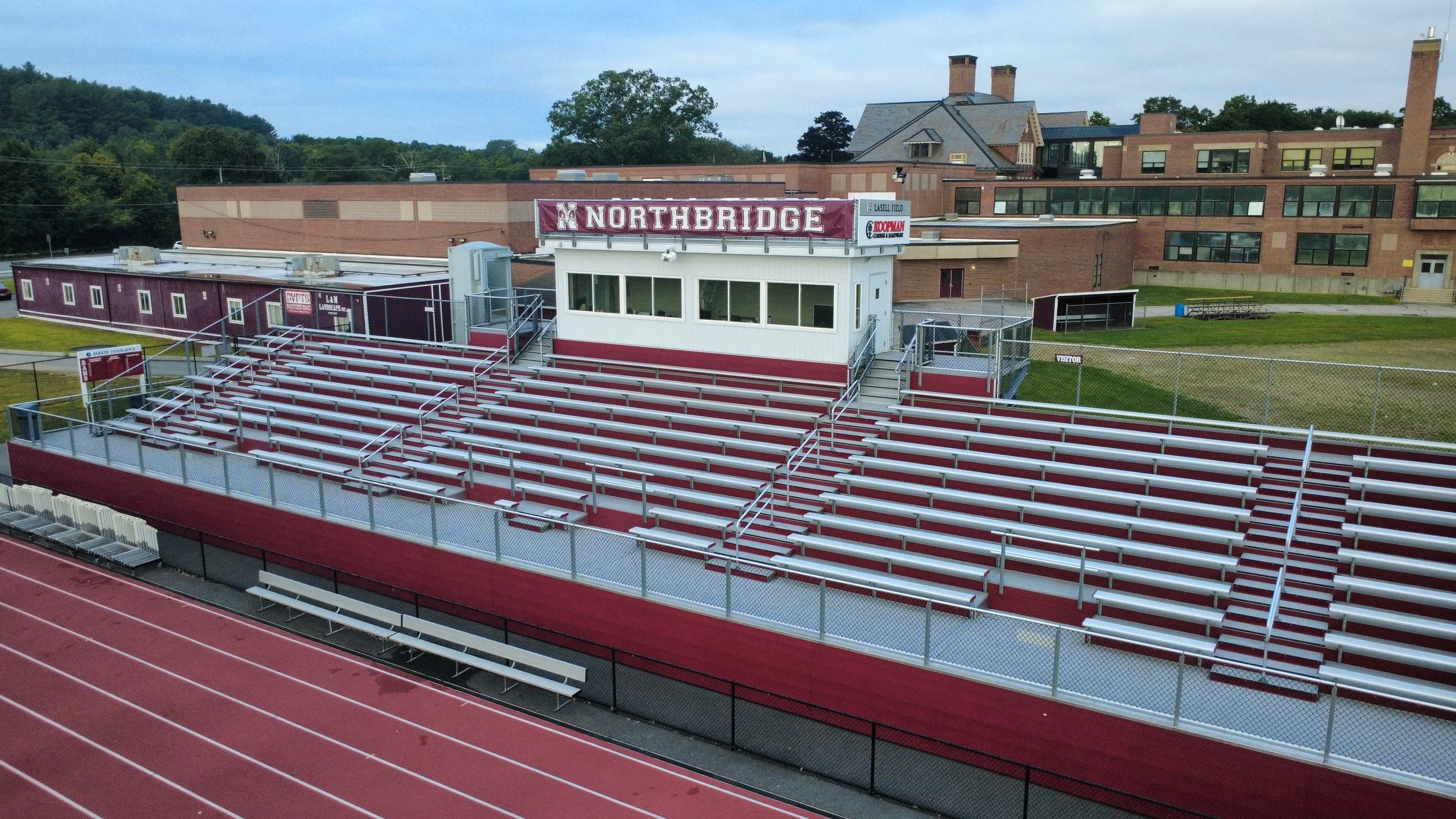 Empty bleachers in a stadium with a white press box featuring a 'NORTHBRIDGE' sign, overlooking a red running track, with school buildings and trees in the background.