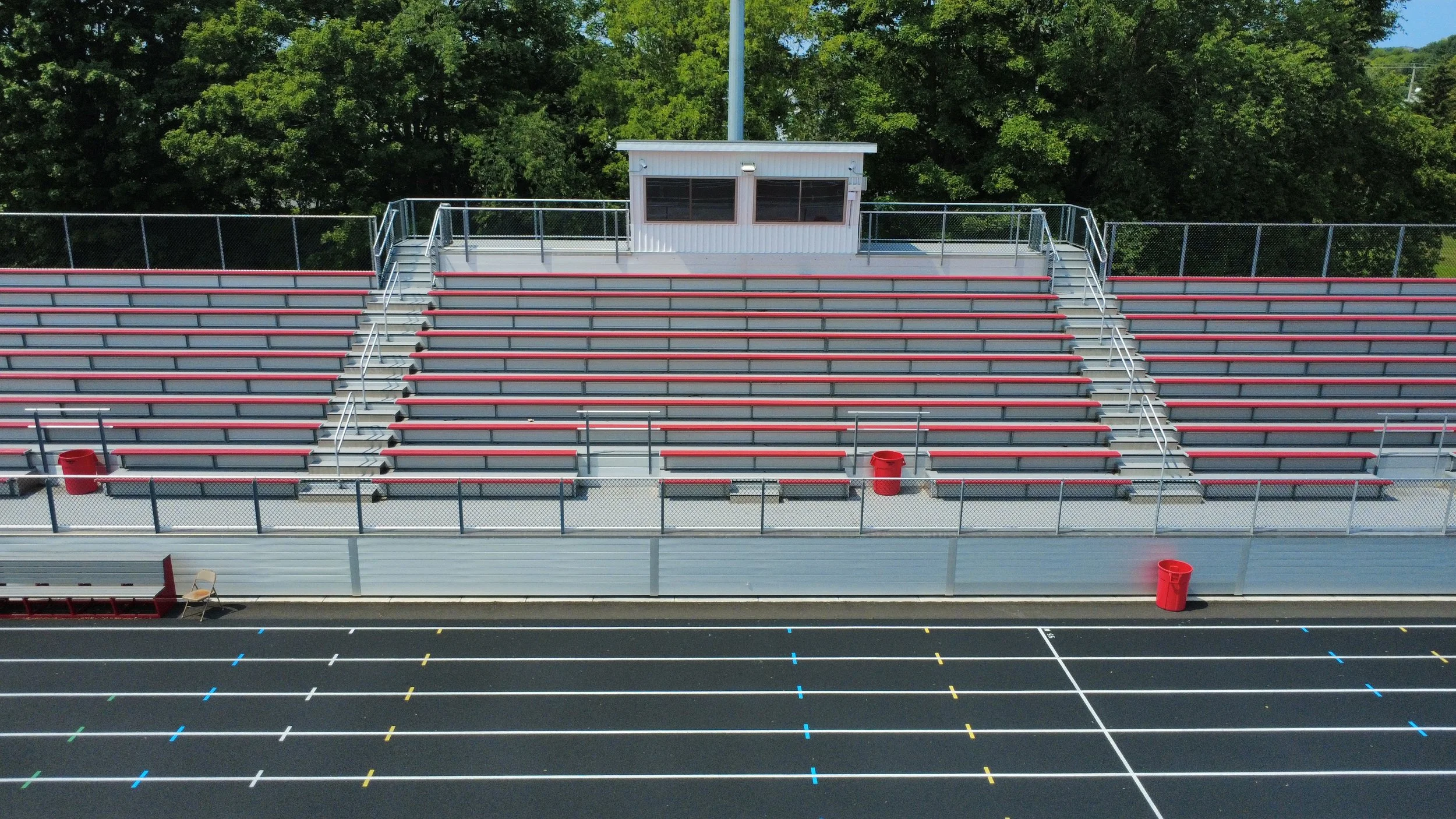 Empty football stadium bleachers with red seats, a small press box at top, and a running track in front with marked lanes. Green trees in background.