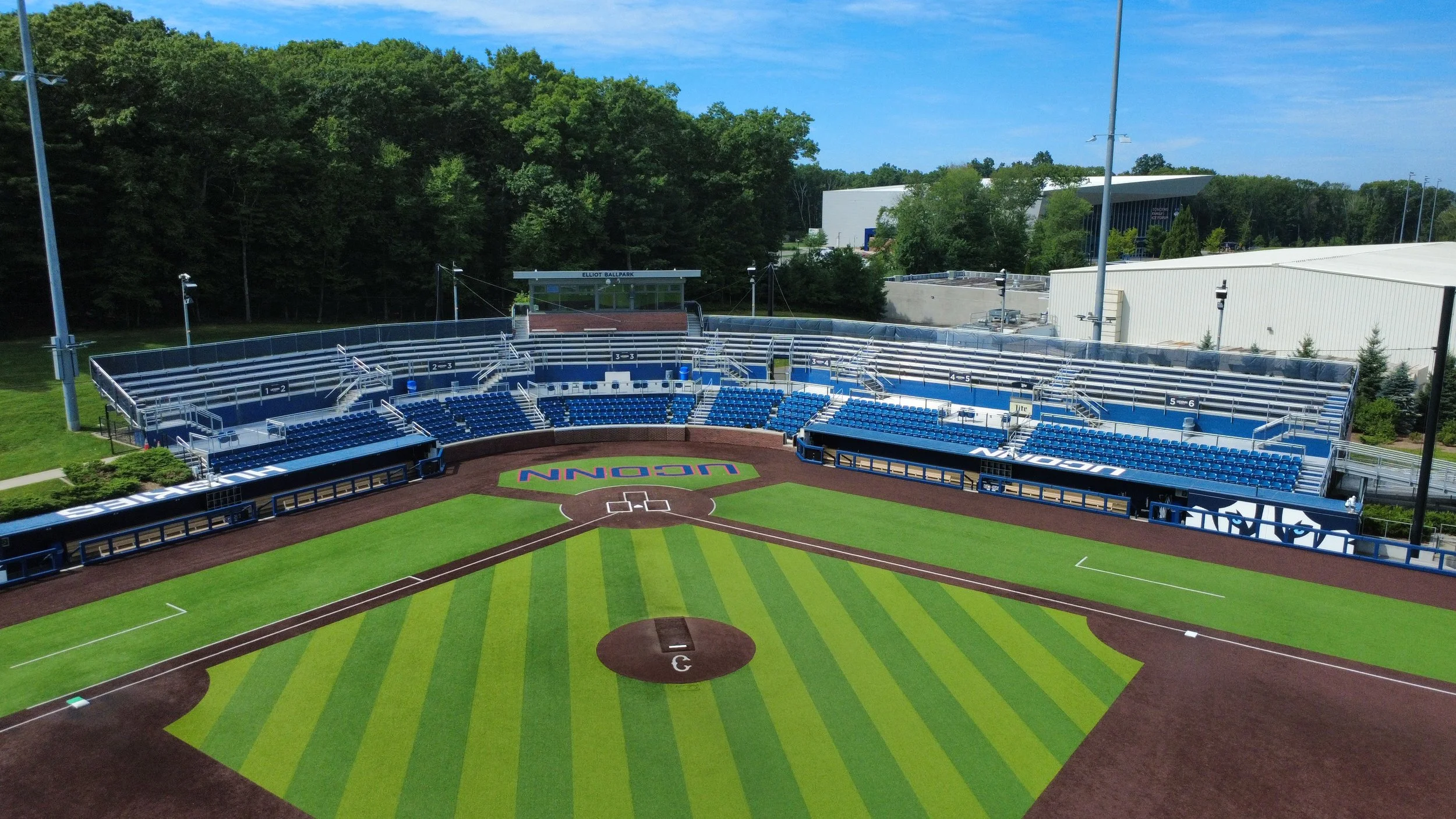 Empty baseball stadium with green grass and a diamond, with blue seating for spectators, surrounded by trees and buildings in the background.