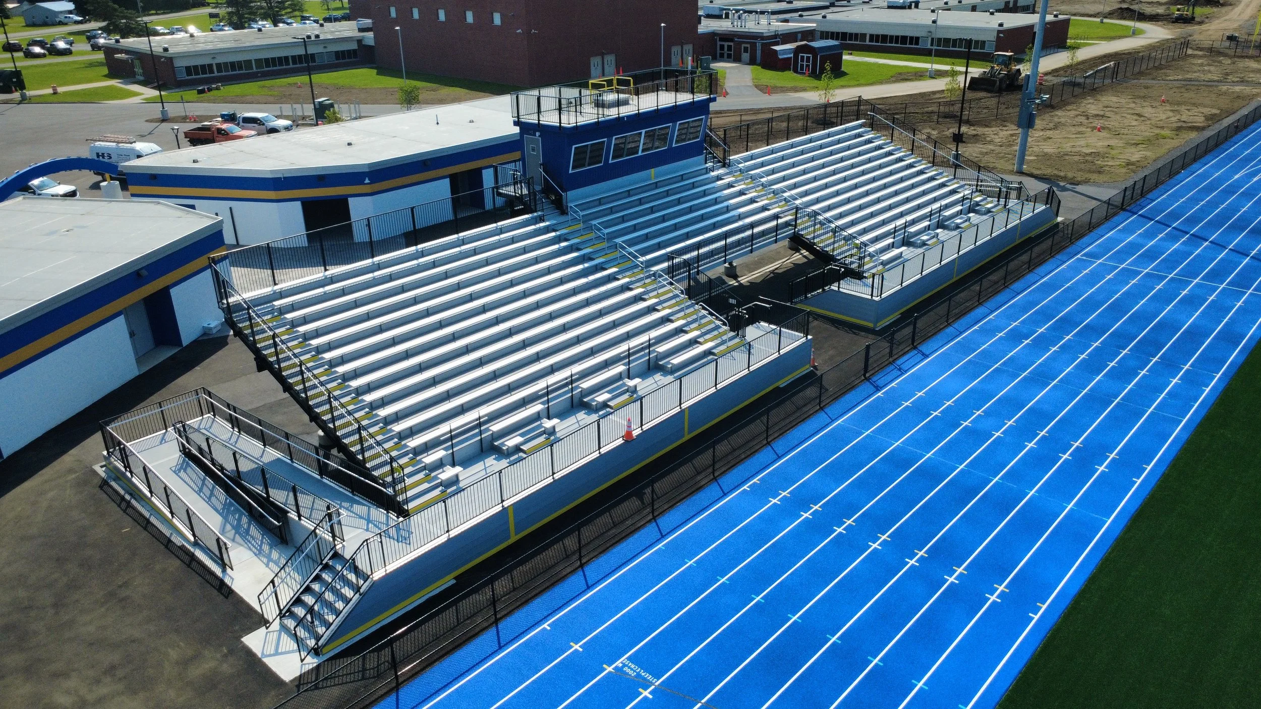 High school football stadium with blue and yellow bleachers, a blue press box, and a blue running track. Surrounding the stadium are parking lots, a school building, and construction areas.