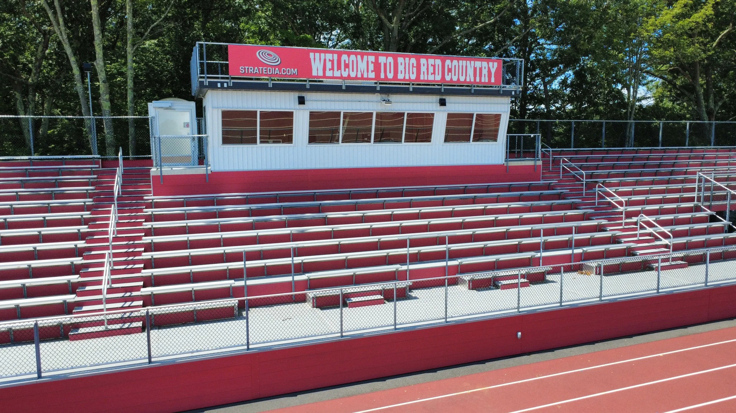 Empty red and silver bleacher seats at an outdoor sports stadium with a small white press box and a large red sign that reads 'Welcome to Big Red Country'.