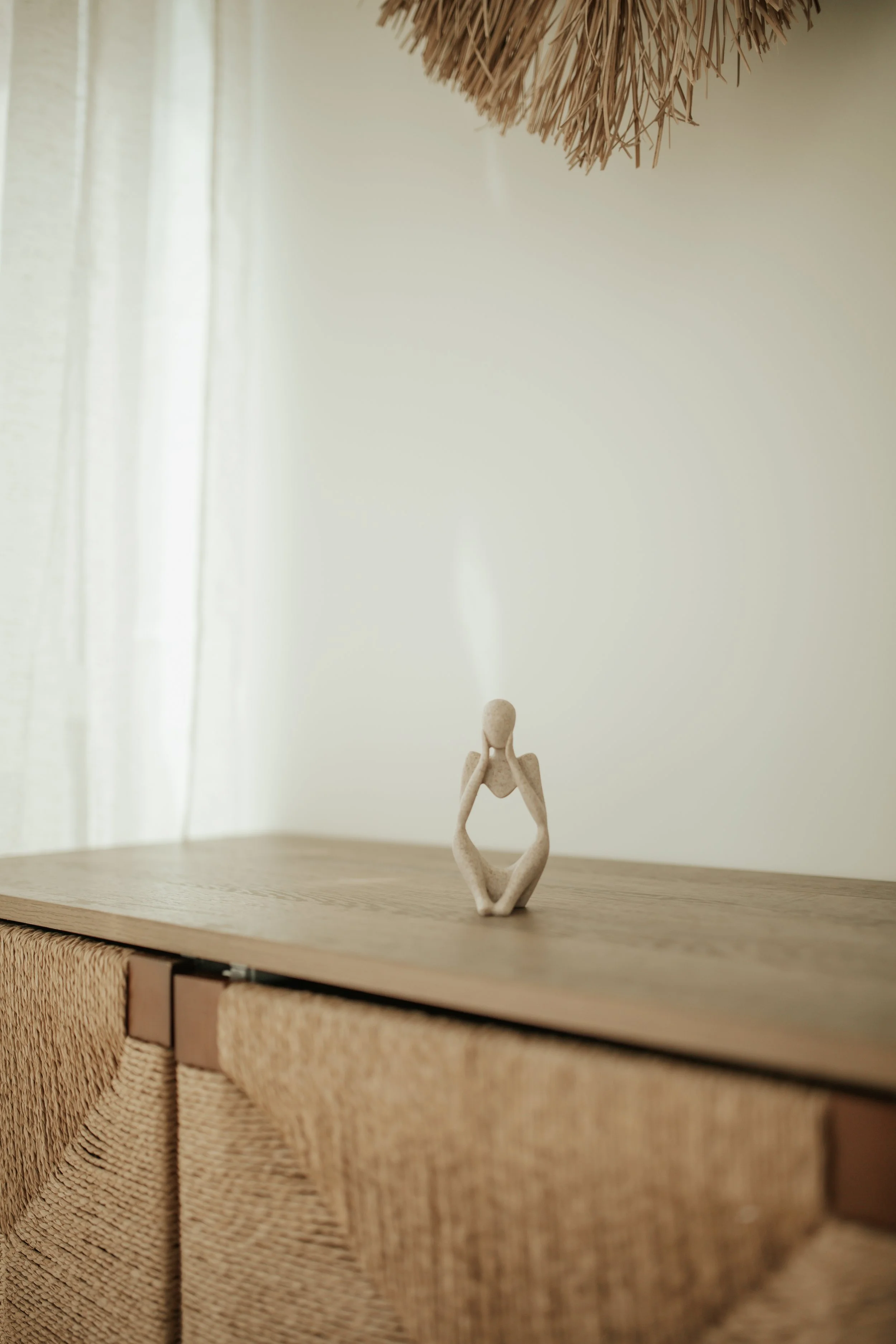 A small, abstract figurine of a person with hands on their face, sitting with knees drawn up, on a wooden surface in a minimalist setting.