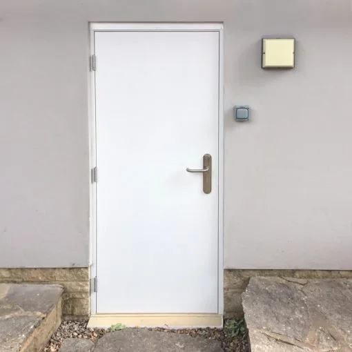 White door with silver handle set into a light gray wall, flanked by two small square light fixtures, with a concrete and stone pathway in front.