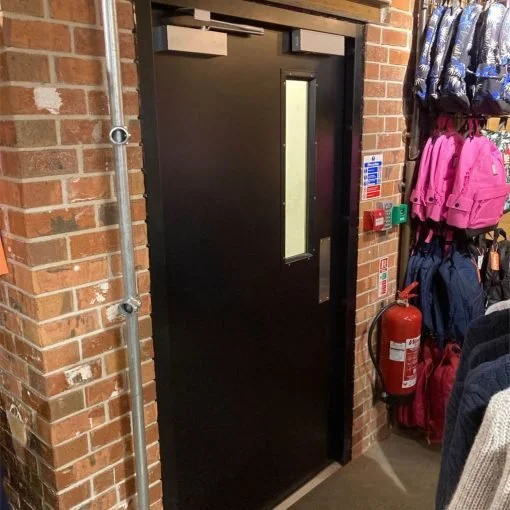 Black emergency exit door with vertical window, brick wall on the left, and colorful backpacks hanging on hooks on the right, with a fire extinguisher and safety signs nearby.