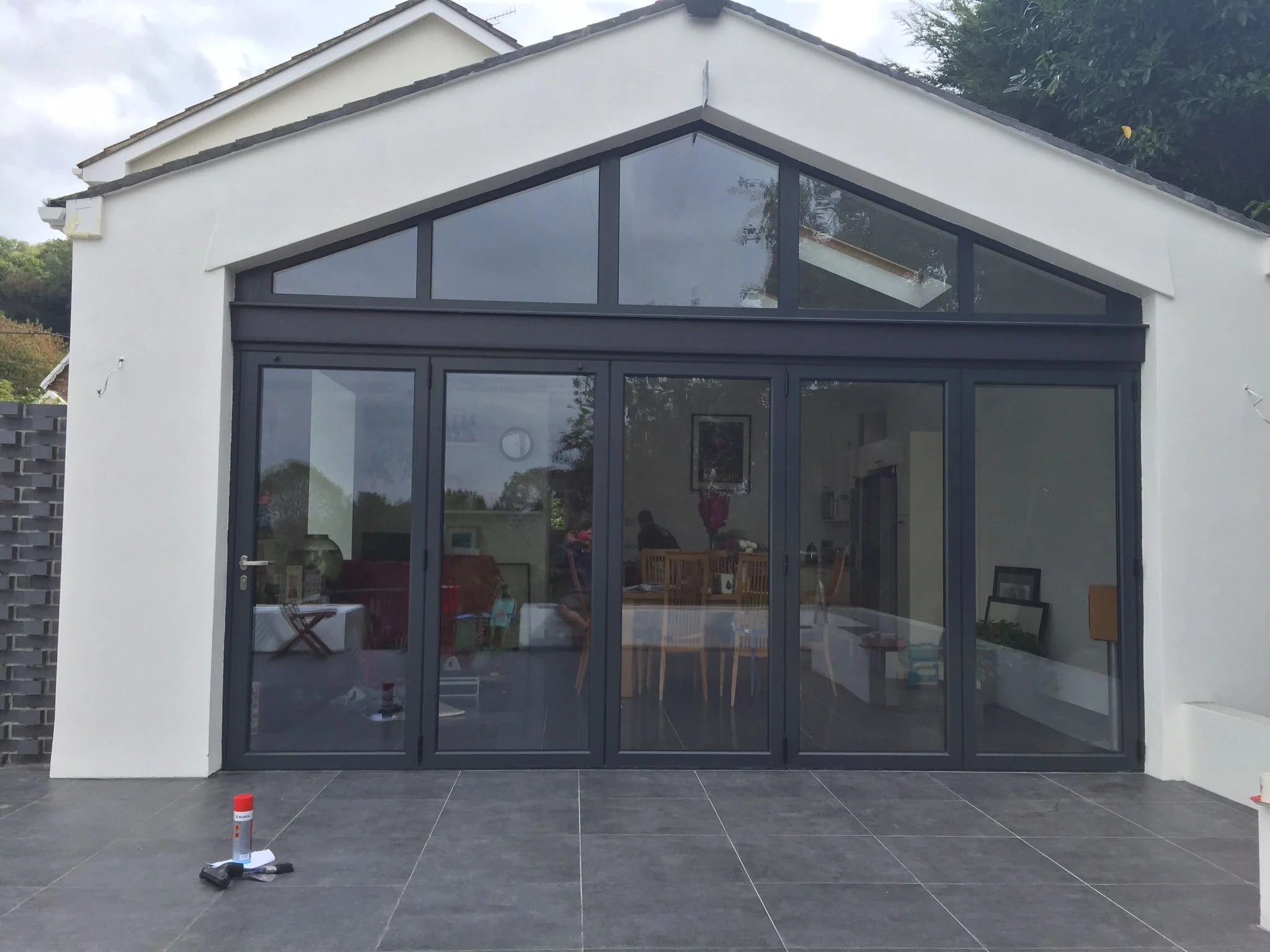 A house with large glass sliding doors and a triangular window above. The exterior features white walls and black window frames. The patio has dark tiles, with some tools and cones lying on the ground.