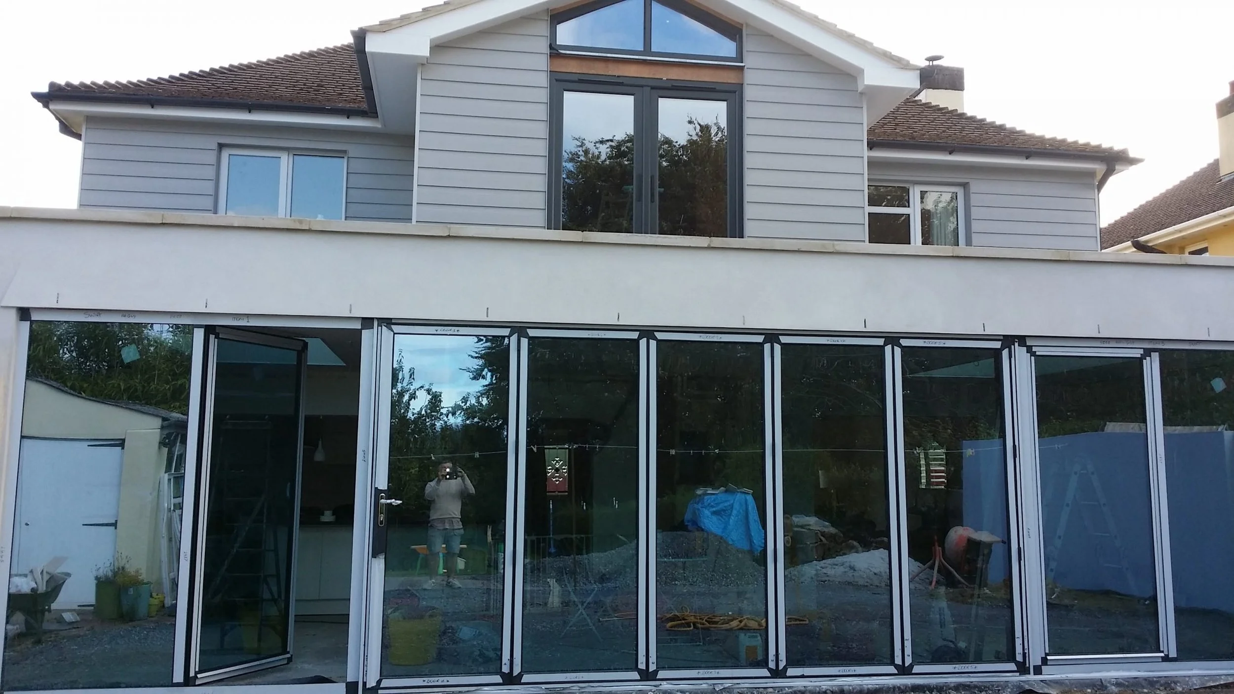 Under construction or renovation of a house, with large glass sliding doors on the ground floor and a second-story room with a balcony and large window, exterior walls painted in light gray, and a tiled roof.