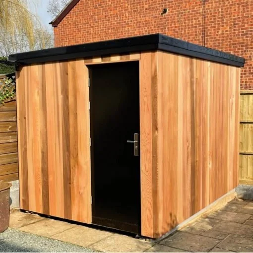 A small wooden storage shed with a black door and black roof, situated in a backyard with a brick wall and a wooden fence.