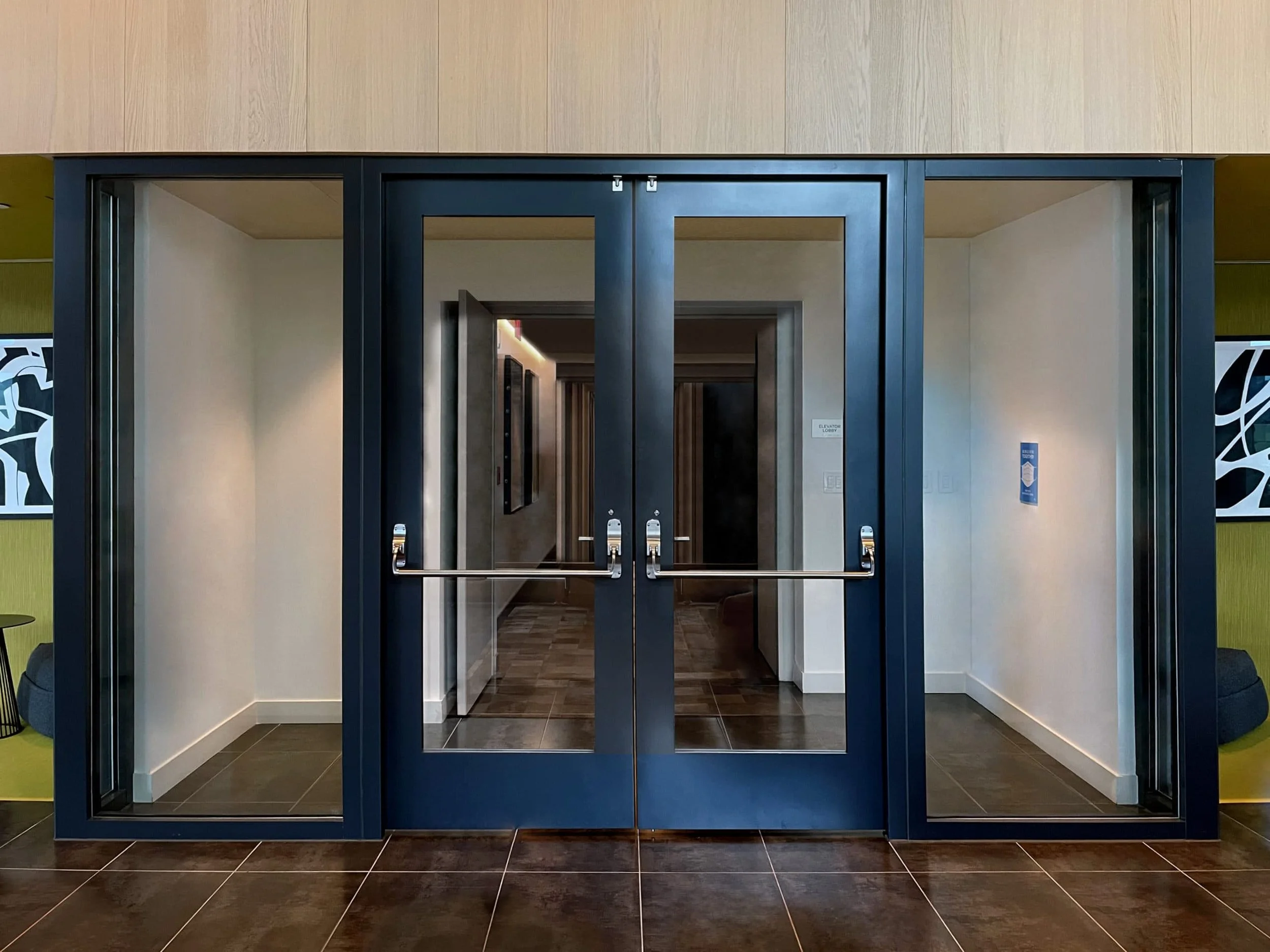 Double glass doors with black frames and automatic door openers in a modern building lobby, showing a hallway with artwork on the walls.