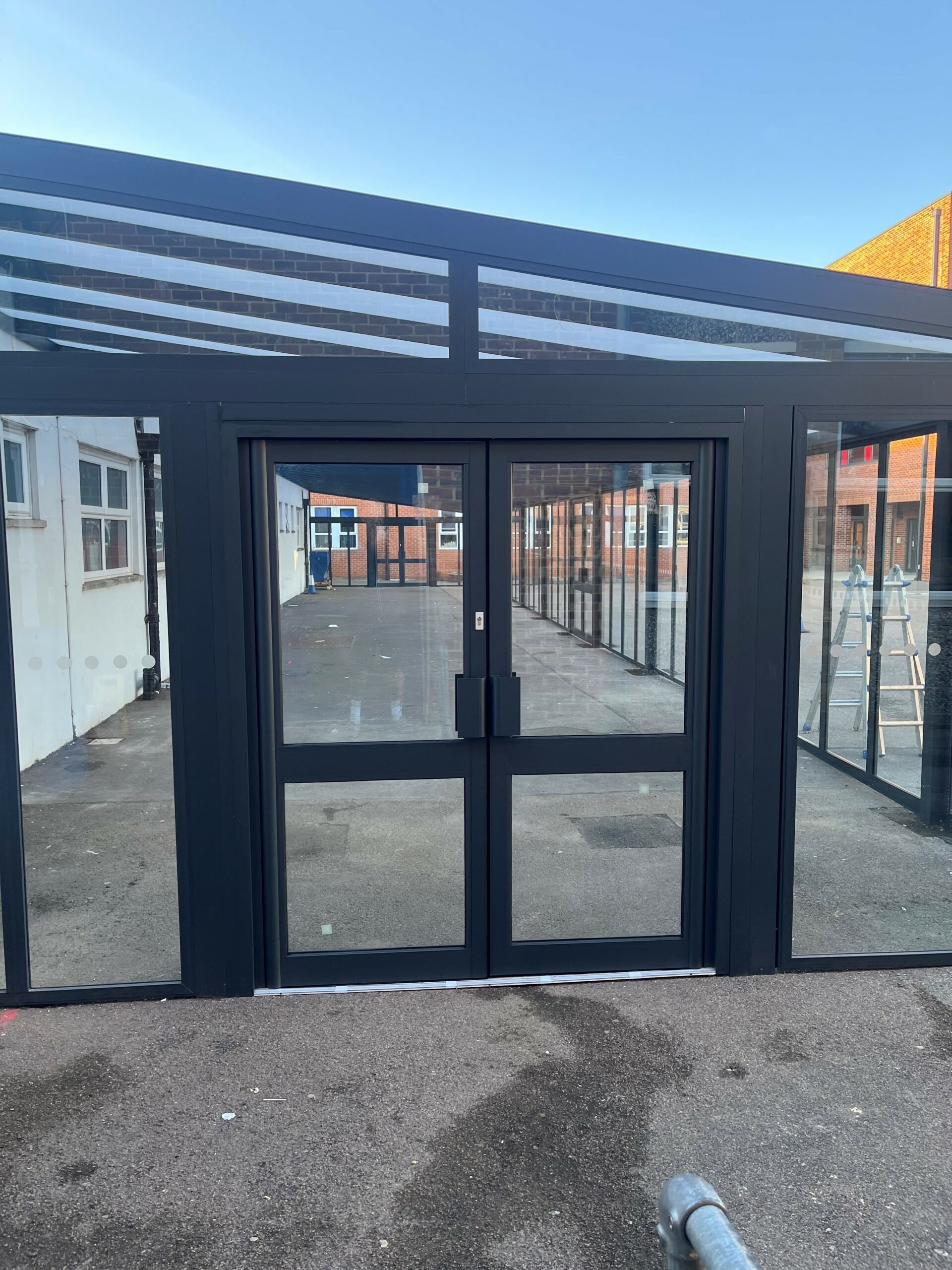 Modern glass-enclosed building entrance with black metal framing and glass doors, surrounded by outdoor pavement and additional glass walls, with a ladder inside and a blue sky above.