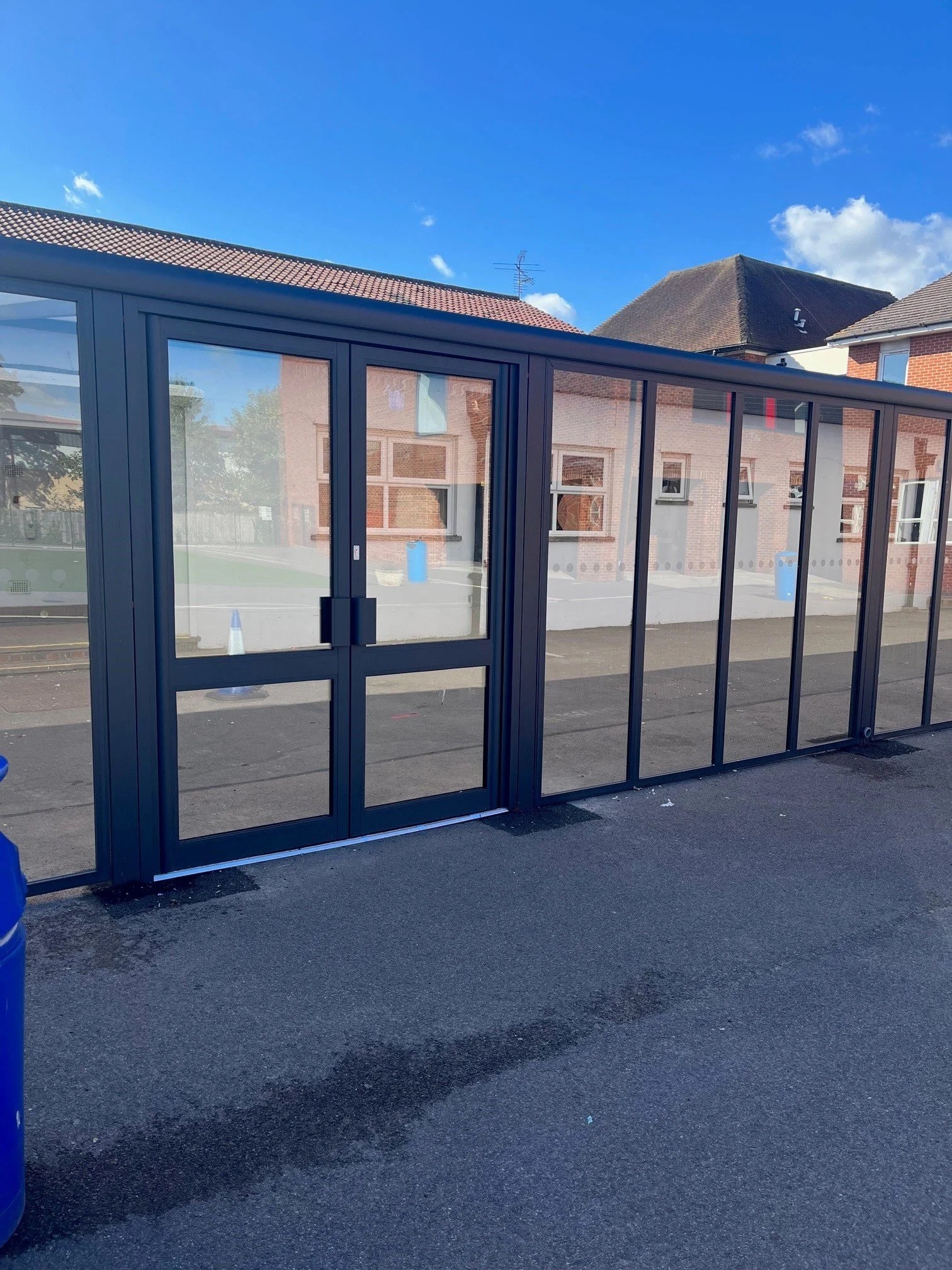 Glass entrance door to a building under construction or renovation, with a double door handle, set in a metal frame, and a partial view of neighboring brick buildings and a clear blue sky.