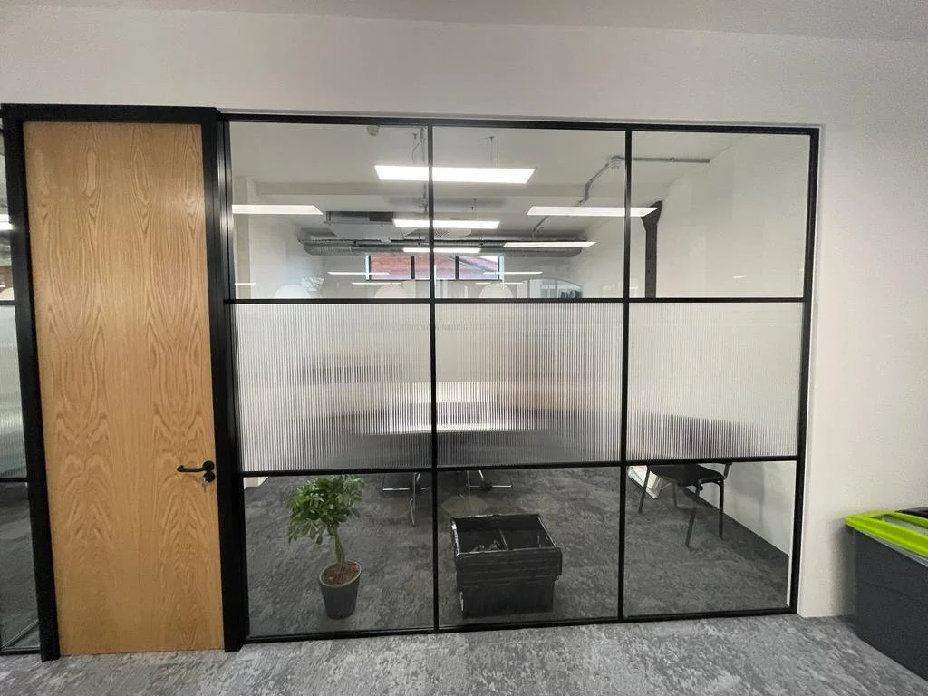 An office conference room with a glass wall, a wooden door, a potted plant, a black trash bin, and a small black chair inside.