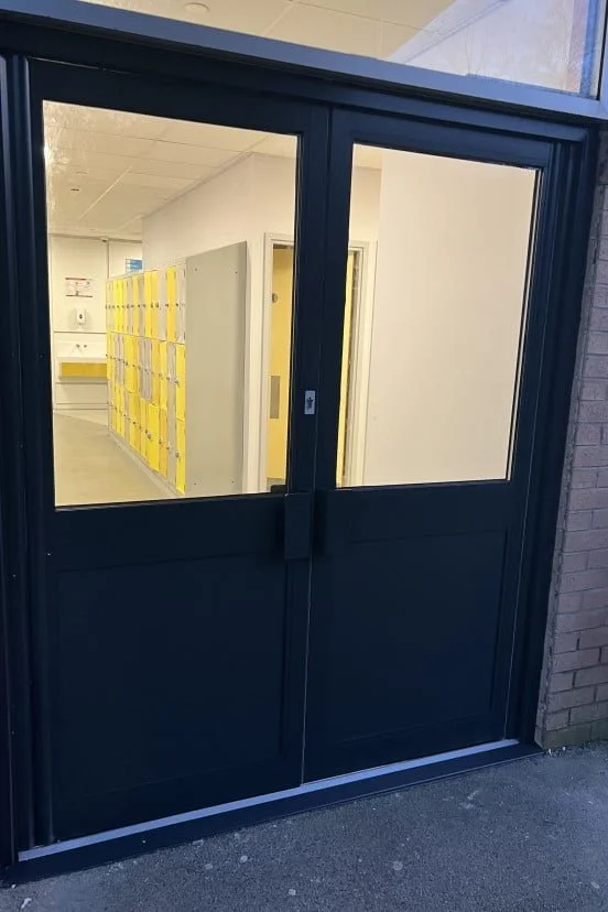Black door with two large glass panels, leading into a hallway with yellow lockers visible inside.
