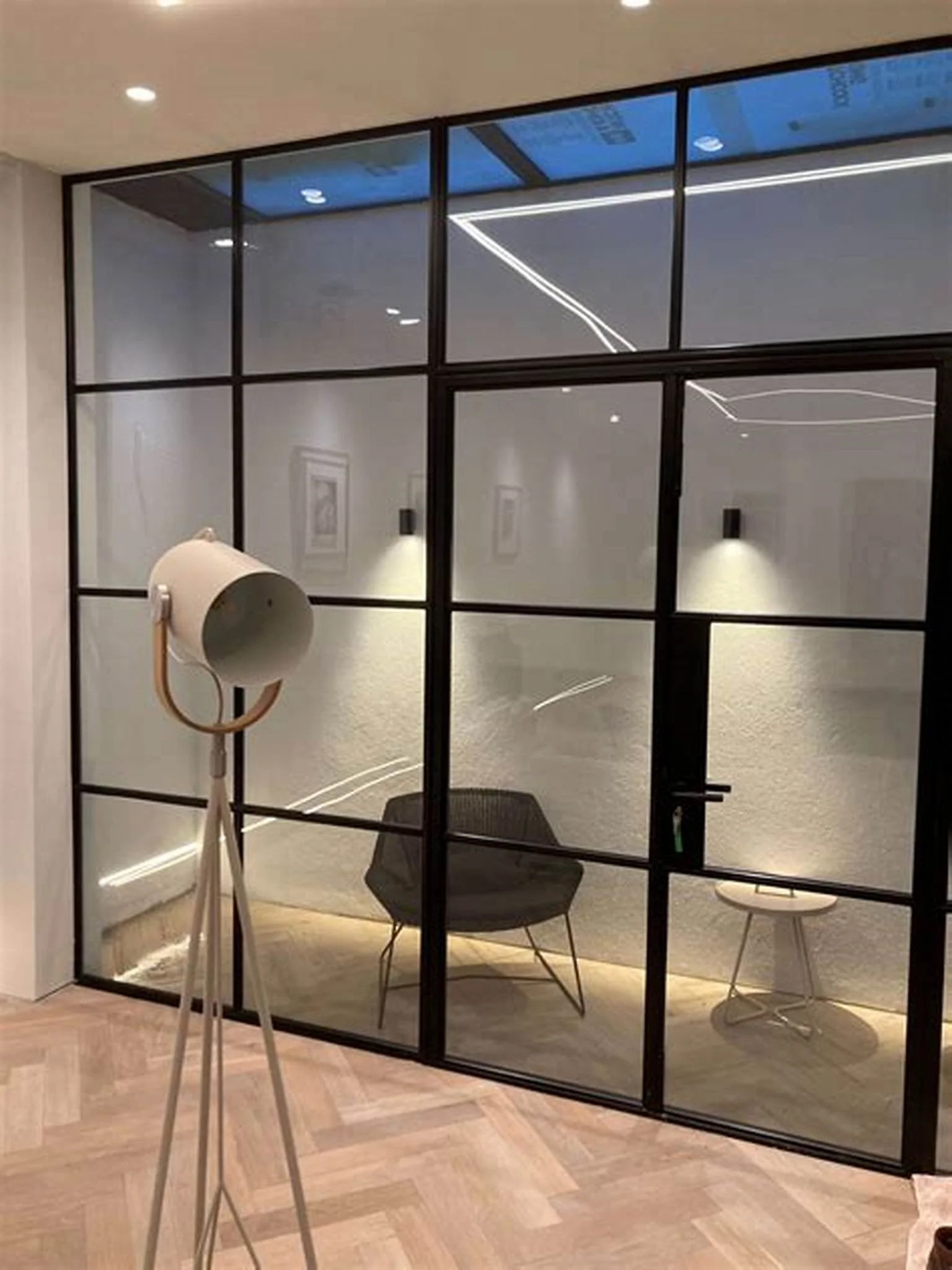 Modern glass-enclosed office with black metal frames, containing a black chair, a small stool, and a standing floor lamp.