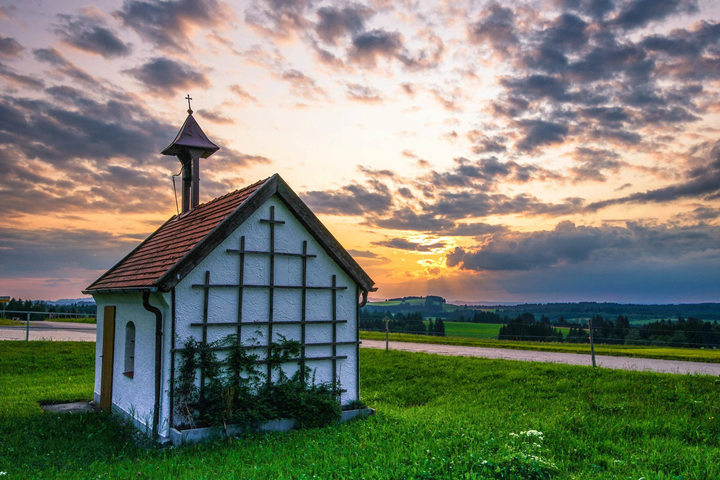 Kleines weißes Kirchengebäude mit rotem Dach vor einem Sonnenuntergang am Himmel, umgeben von grüner Wiese und offenen Feldern.