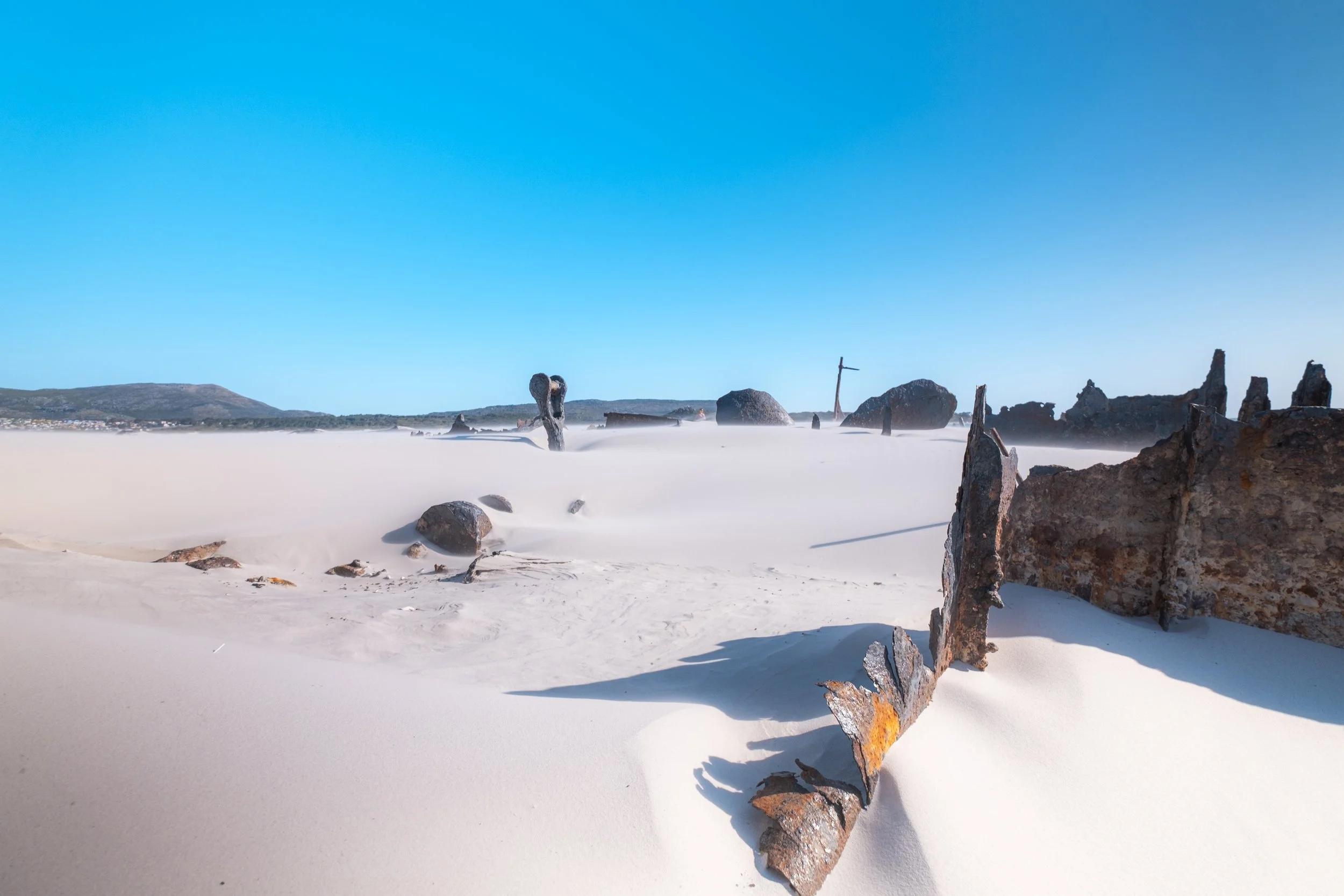 Sandstrand mit rostigen Metallresten und großen Steinen, blauer Himmel, ferne Berge im Hintergrund.