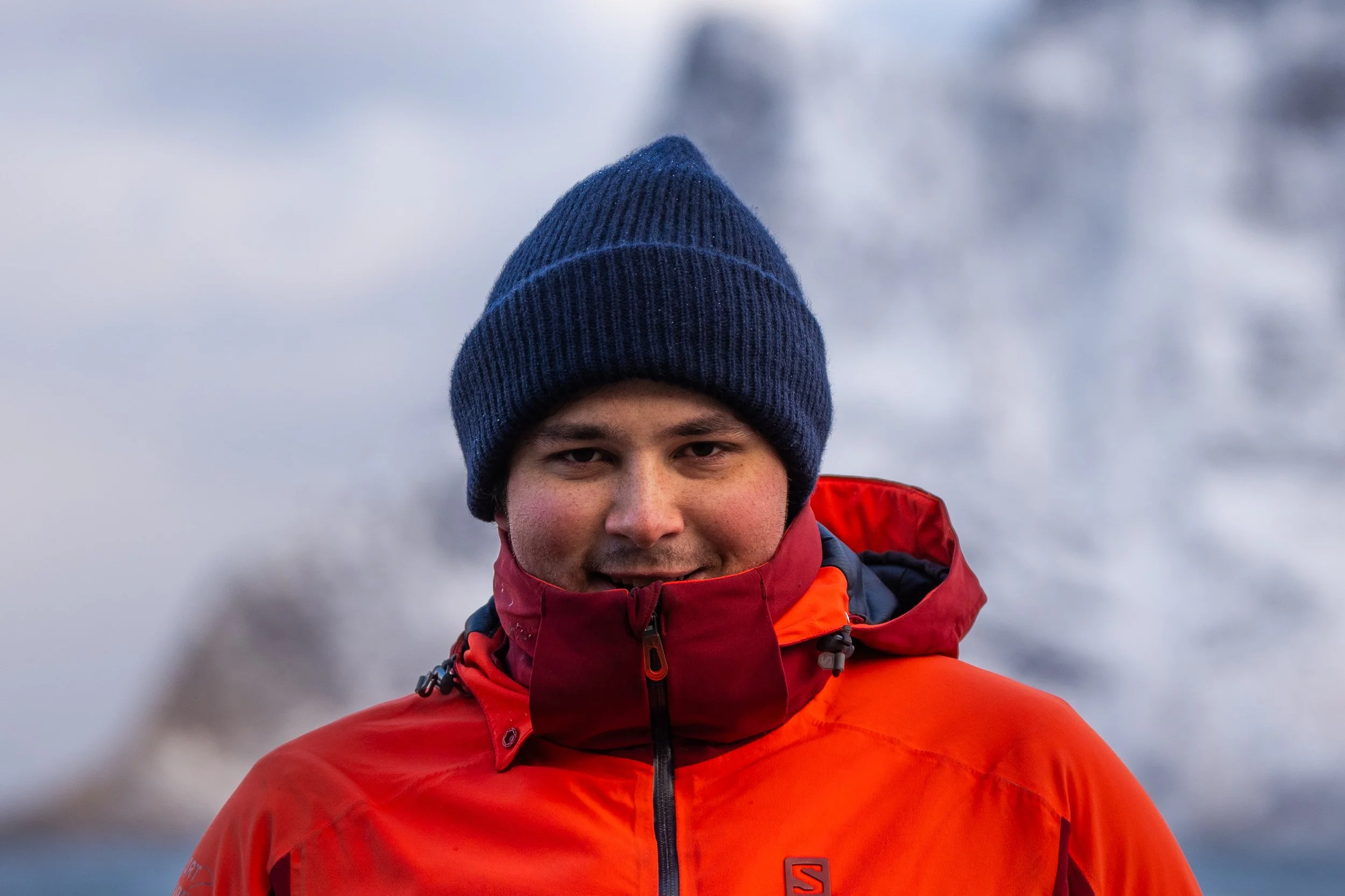 Junge Person in roter Jacke und blauer Mütze vor verschneiter Berglandschaft.