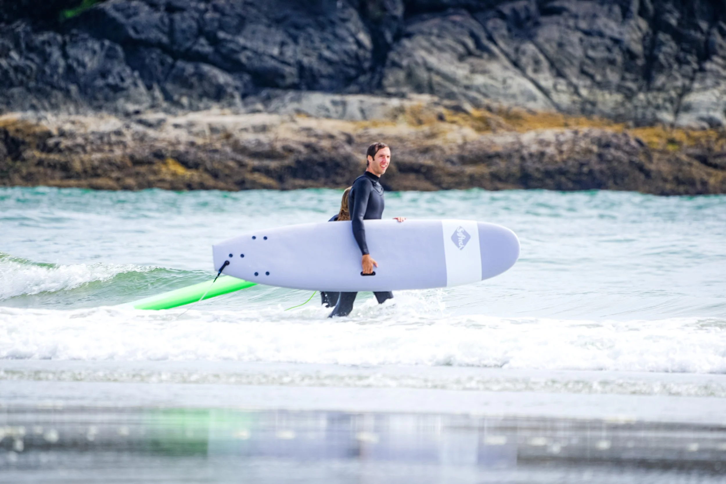 Mann beim Surfen im Wasser, hält Surfbrett in der Hand, trägt Neoprenanzug, im Hintergrund felsige Küste.