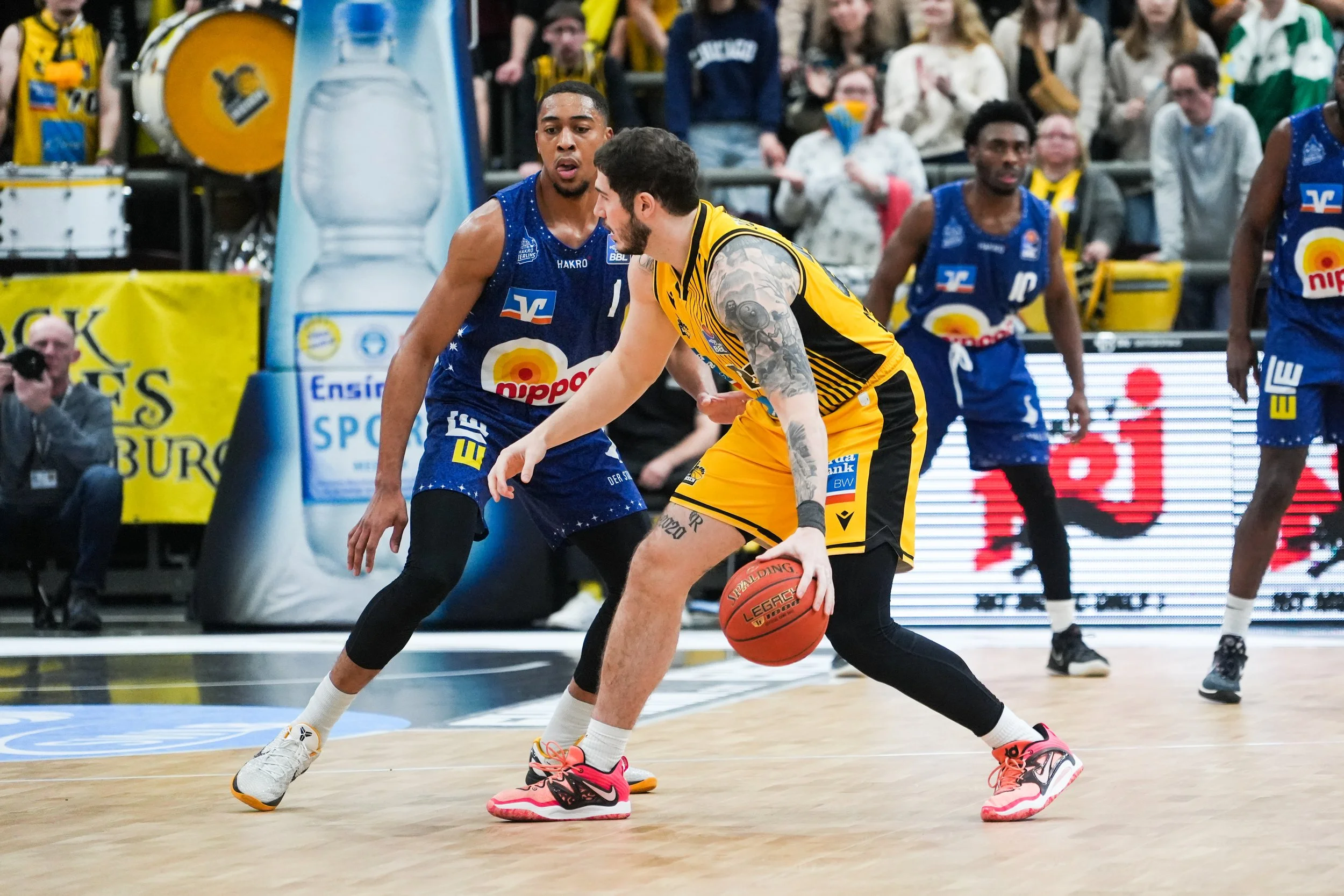 Basketballspieler in Aktion während eines Spiels, einer in gelbem Trikot, der andere in blauer Trikot, auf einem Basketballfeld mit Publikum im Hintergrund.