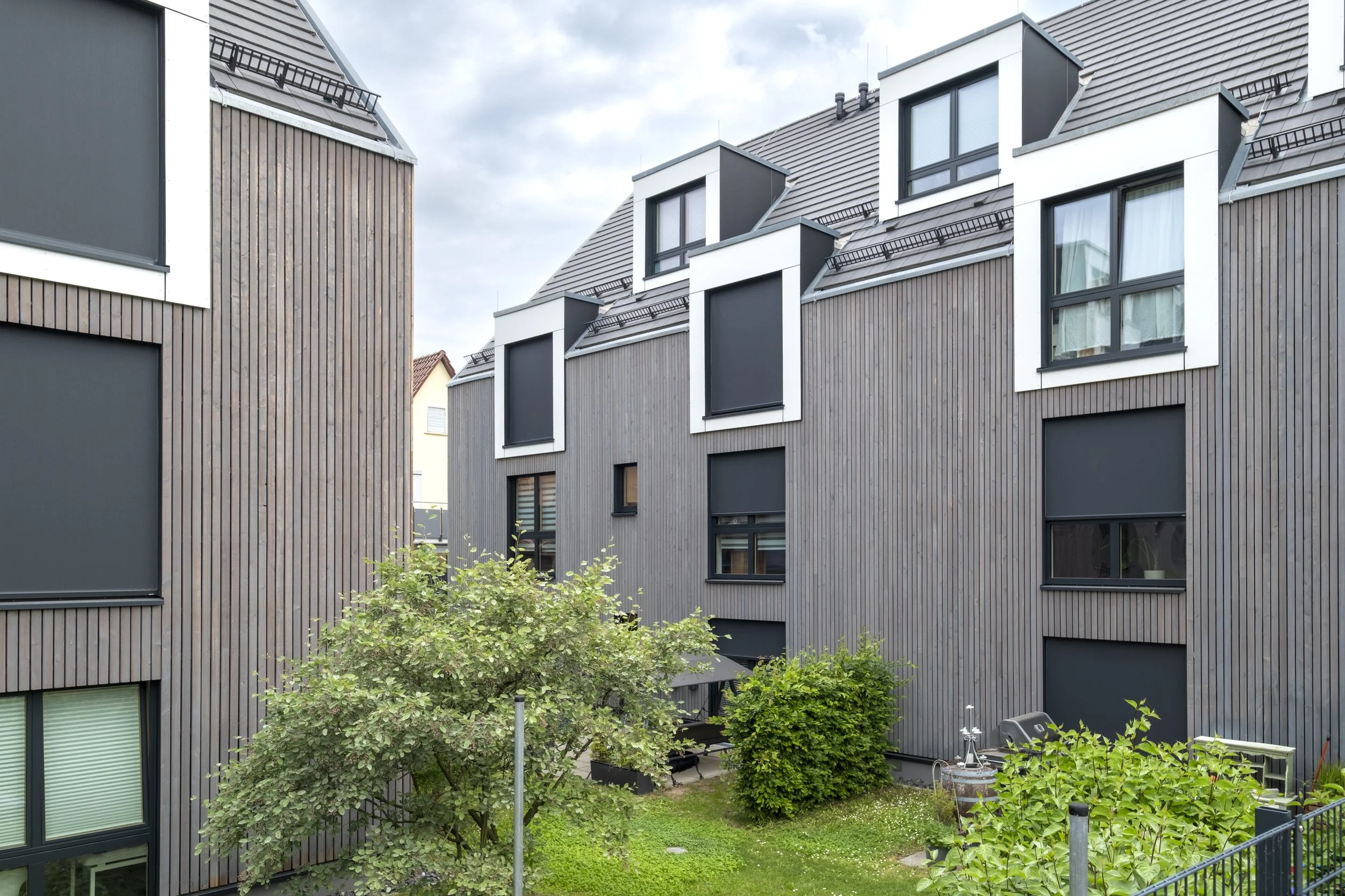 Moderne Mehrfamilienhäuser mit Holzfassade, große Fenster mit Rollläden, kleiner Garten mit Bäumen und Bügeln im Innenhof