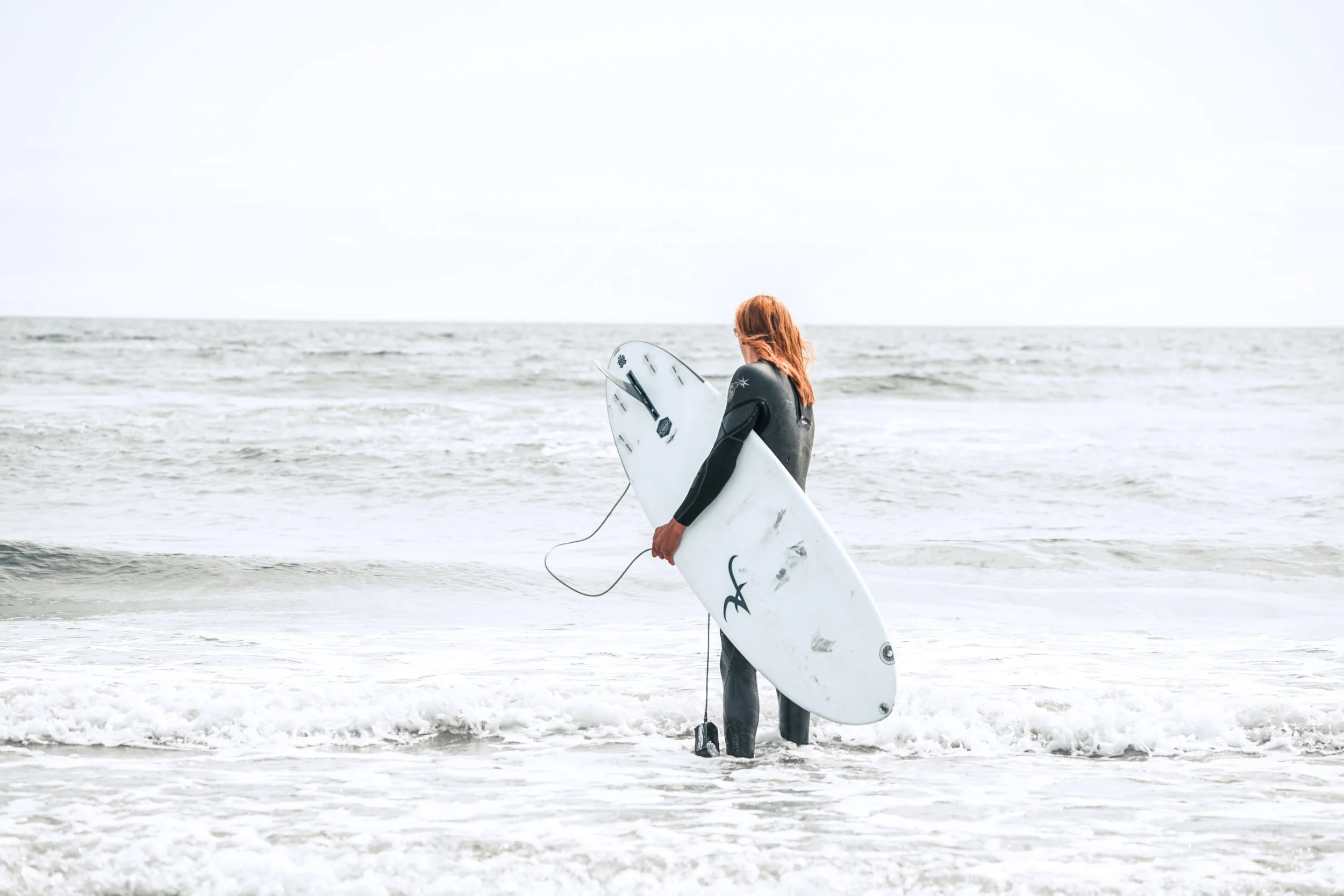 Surferin mit rotem Haar, schwarze Neoprenanzug, läuft ins Meer mit einem weißen Surfbrett mit schwarzen Markierungen