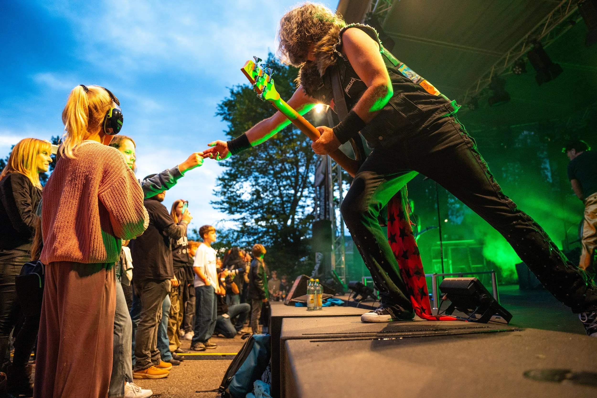 Ein Gitarrist im schwarzen Outfit mit Konfetti-Design fordert ein Publikum, das auf einer Festival-Bühne steht, hervor. Die Szene ist bei Dämmerung mit buntem Bühnenlicht und einem großen Baum im Hintergrund.