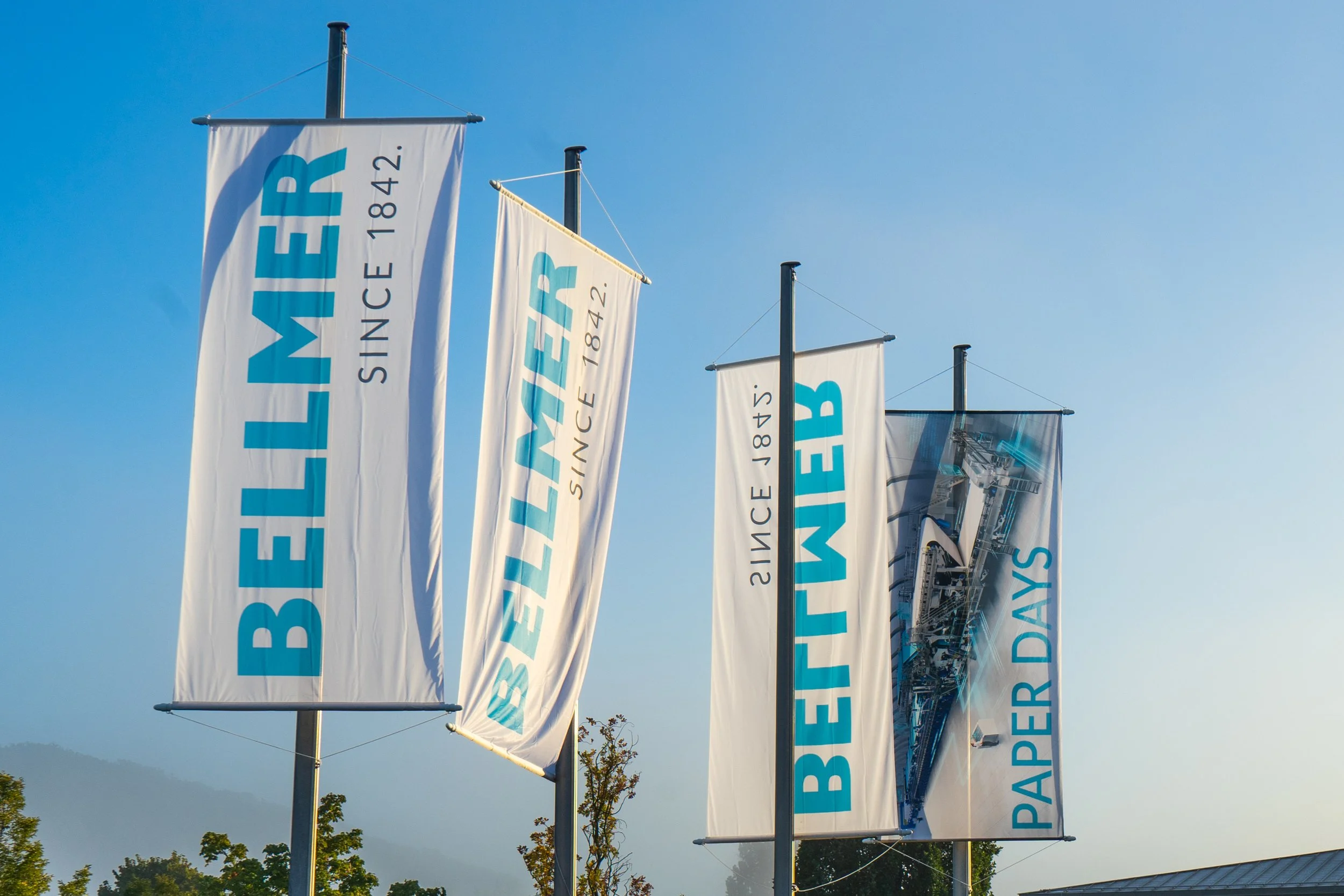 Vier vertikale Fahnen mit dem Text 'BEMMER' und 'SINCE 1842', eine Fahne mit einem technischen Druck oder einer Maschine, und eine weitere Fahne mit dem Text 'PAPER DAYS' gegen einen blauen Himmel.