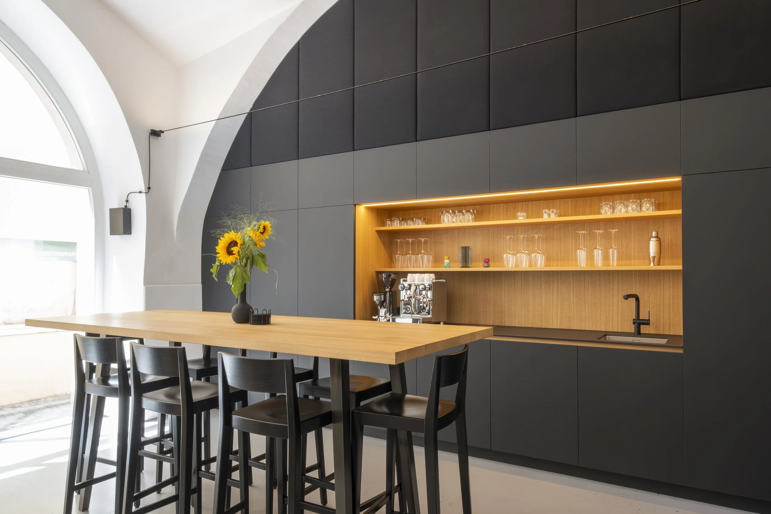 Moderne Bar mit schwarzer und holzfarbener Einrichtung, Sonnenblumen in Vase auf Holztisch, offener Raum mit natürlichem Licht.