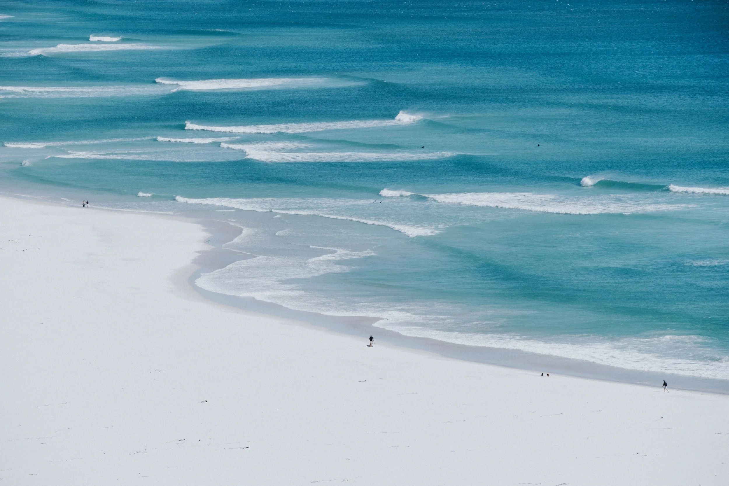 Strand mit weißem Sand, ruhige Wellen im türkisblauen Meer, wenige Menschen spazieren entlang des Ufers, sonniges Wetter.