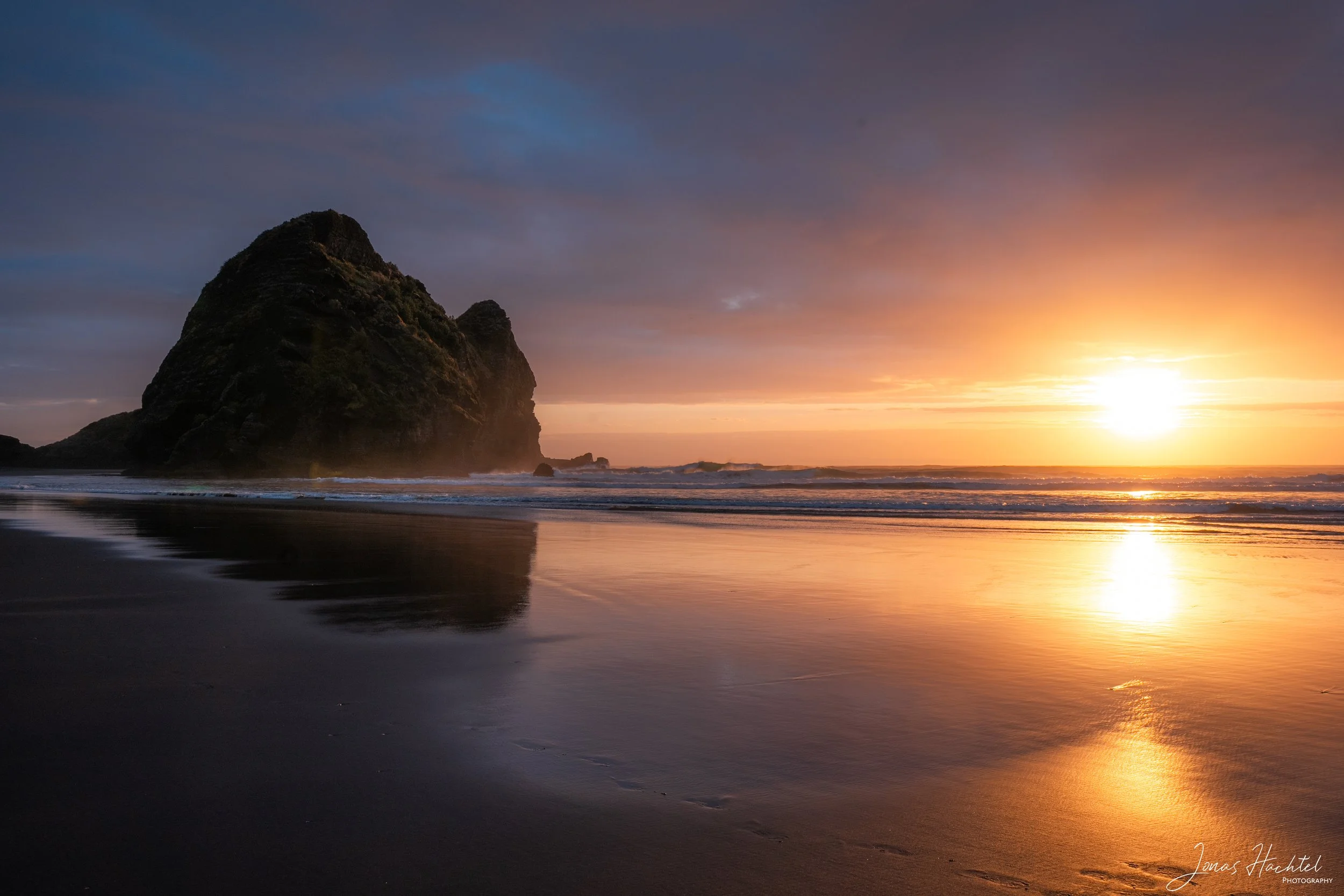Sonnenuntergang am Strand mit großen Felsen im Wasser und Reflexionen auf nassem Sand.