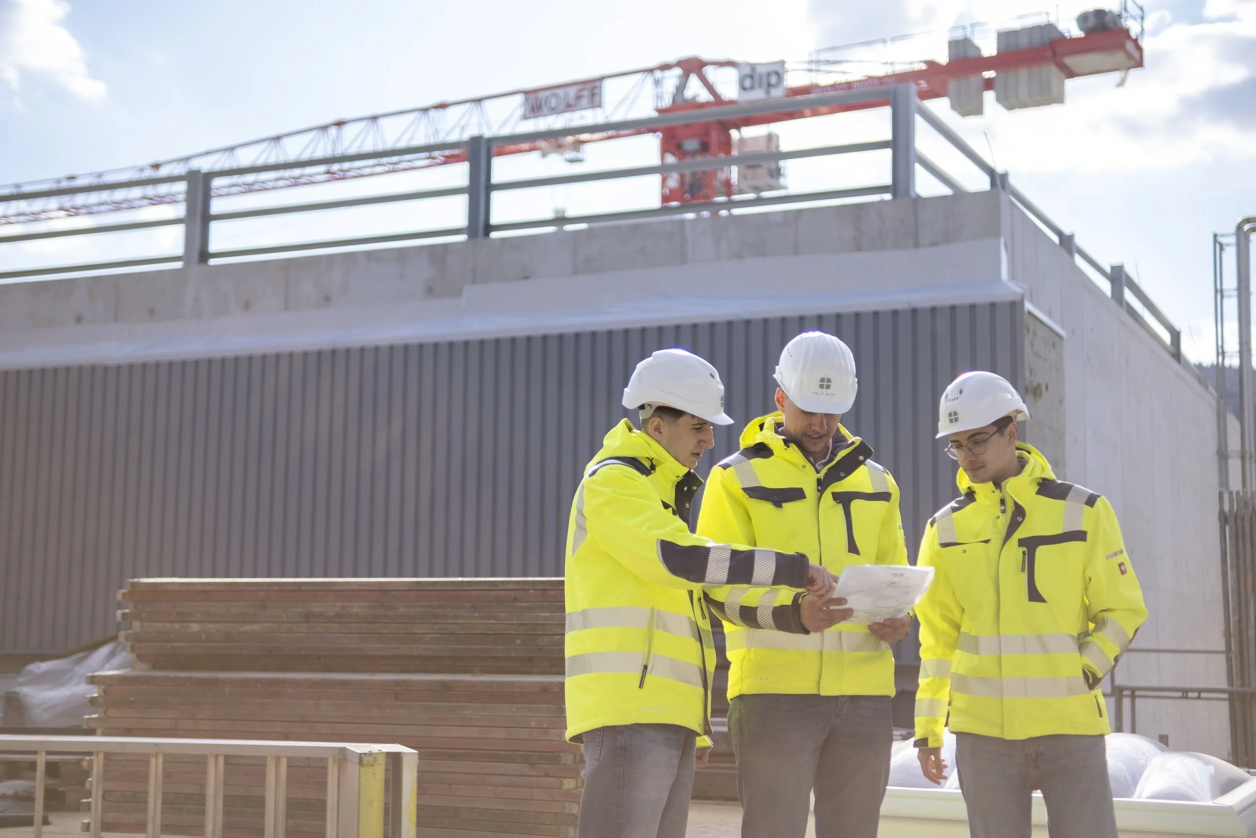 Drei Bauingenieure in gelben Sicherheitsjacken und Helmen diskutieren auf einer Baustelle, während sie einen Plan betrachten.