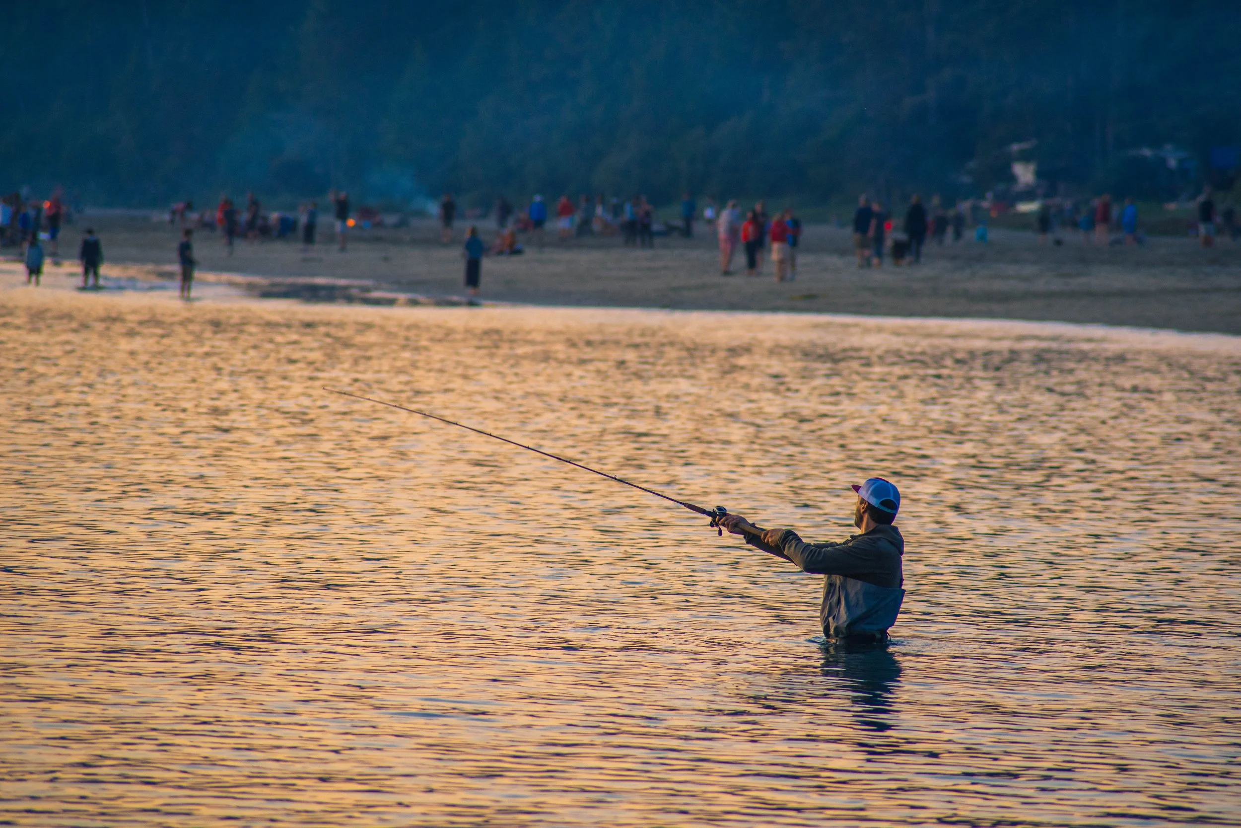 Mann steht im Wasser und fischt während des Sonnenuntergangs, im Hintergrund sind viele Menschen am Strand.