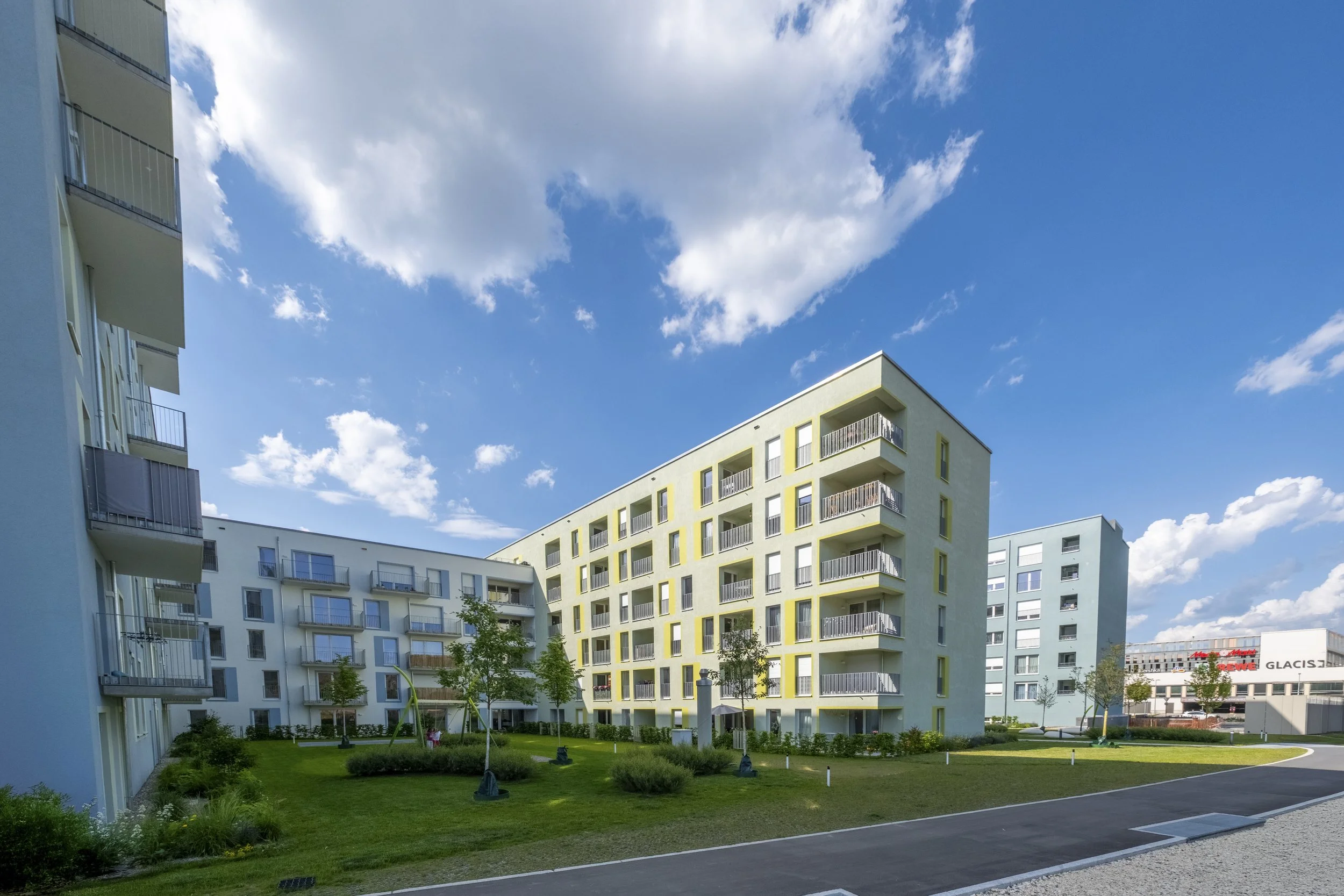 Mehrstöckige Wohngebäude mit Balkon vor einem blauen Himmel mit Wolken, umgeben von Grünflächen und Bäumen.