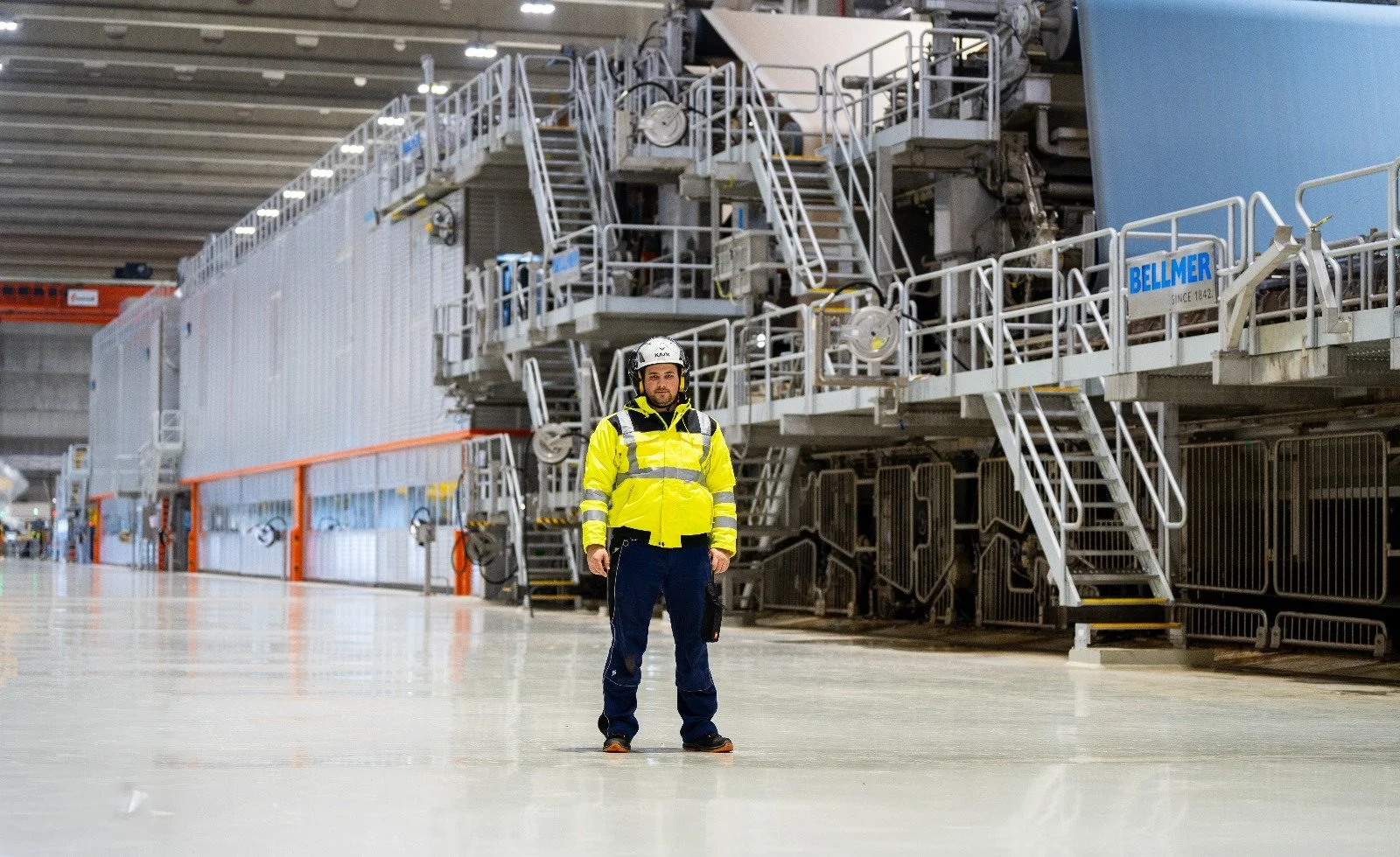 Ein Mann in Hochsicht, gelber Sicherheitsjacke und Helm steht in einer großen Industriehalle vor einer großen Maschine.