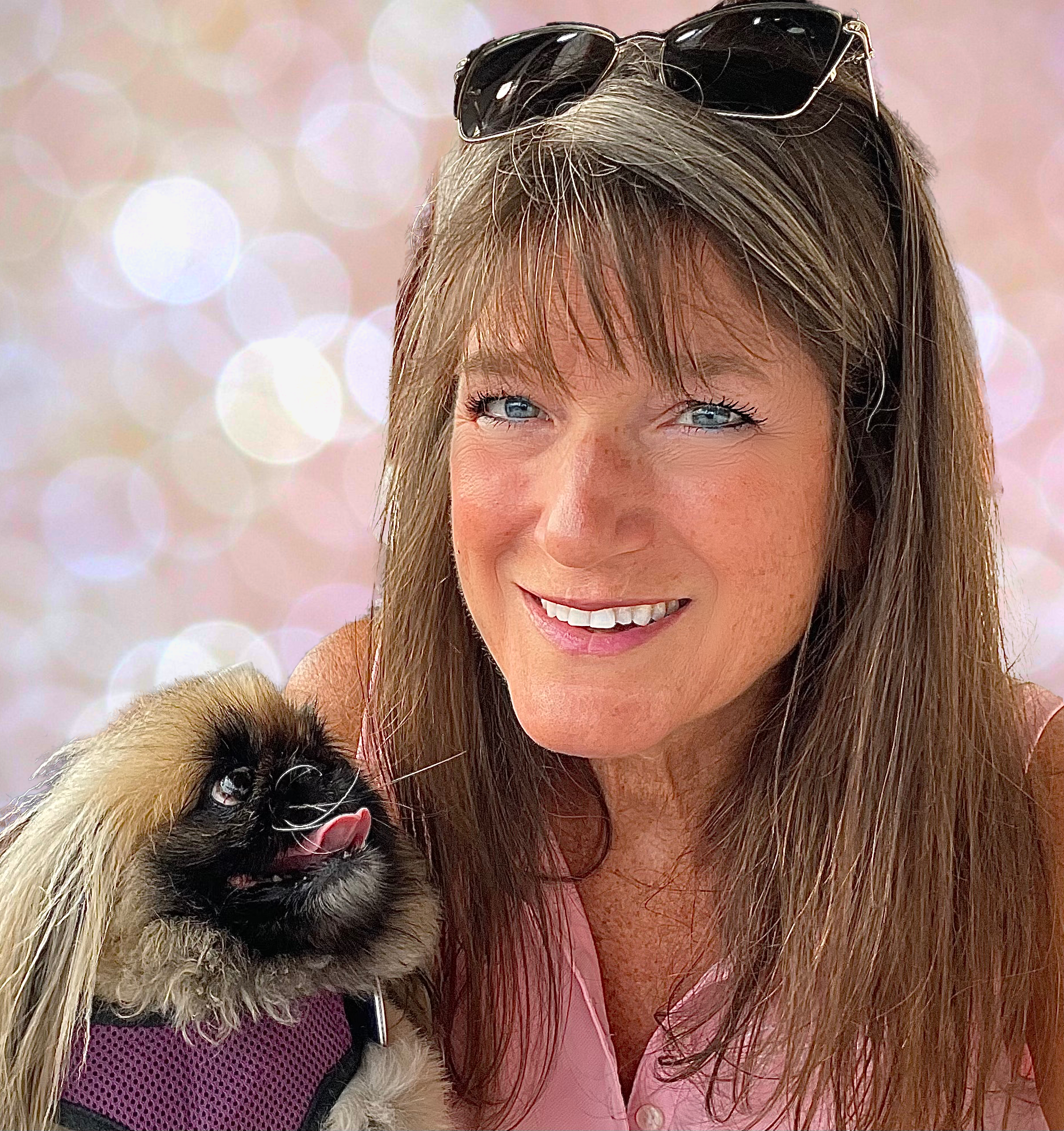 A woman with long brown hair and sunglasses on her head smiling while holding a small dog with long ears and black and beige fur, against a pinkish bokeh background.