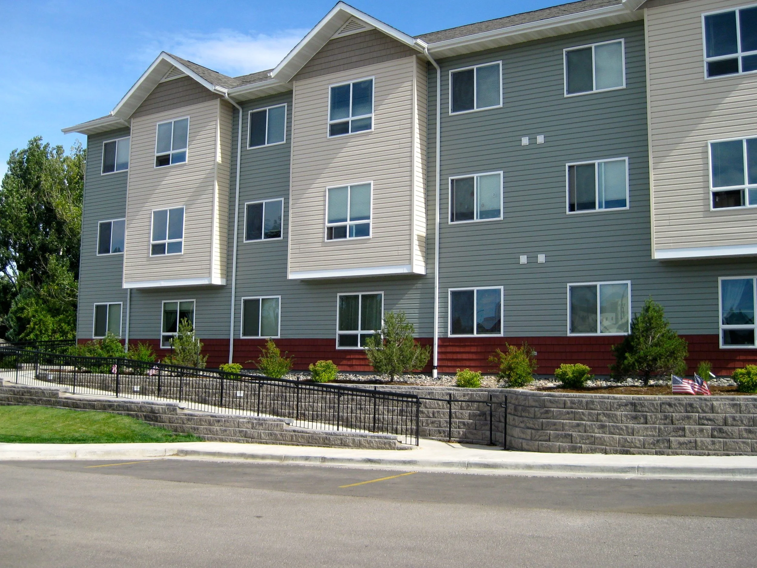 Exterior of a multi-story residential building with beige and green siding, multiple windows, a landscaped area with bushes and small trees, a black metal fence, and a curved stone retaining wall beside a parking lot.