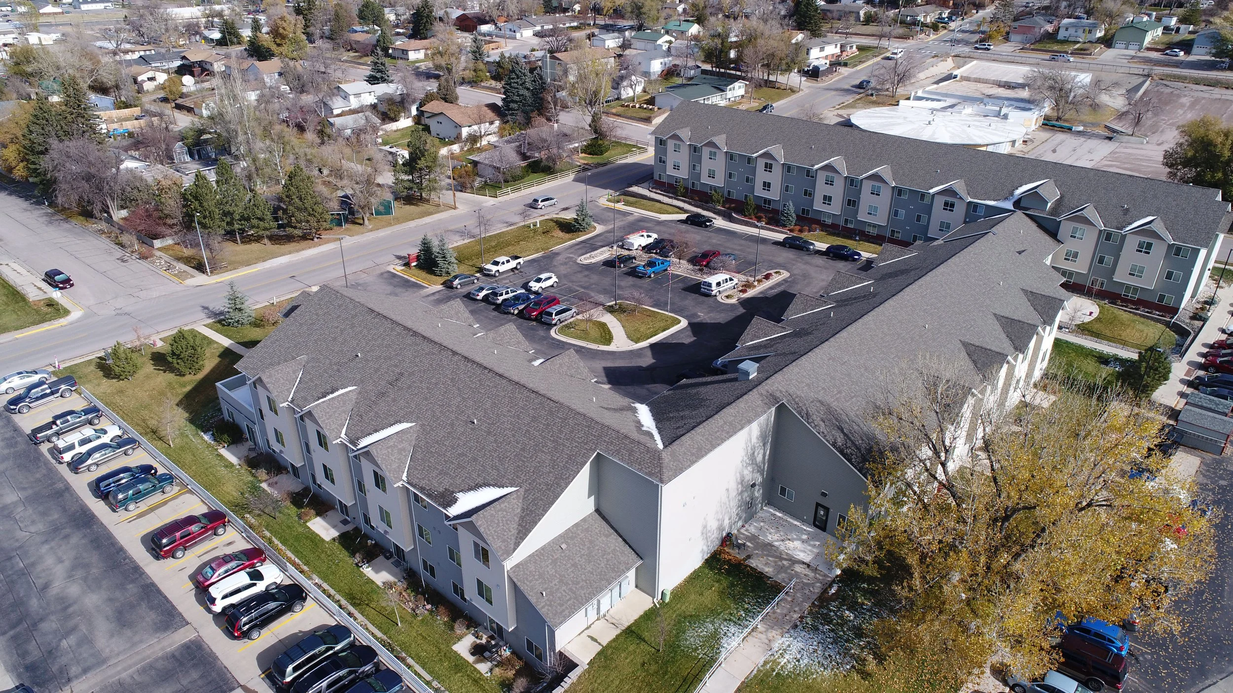 Aerial view of a residential complex with multiple buildings, a parking lot, surrounding houses, and trees. Some snow patches are visible on rooftops and ground.
