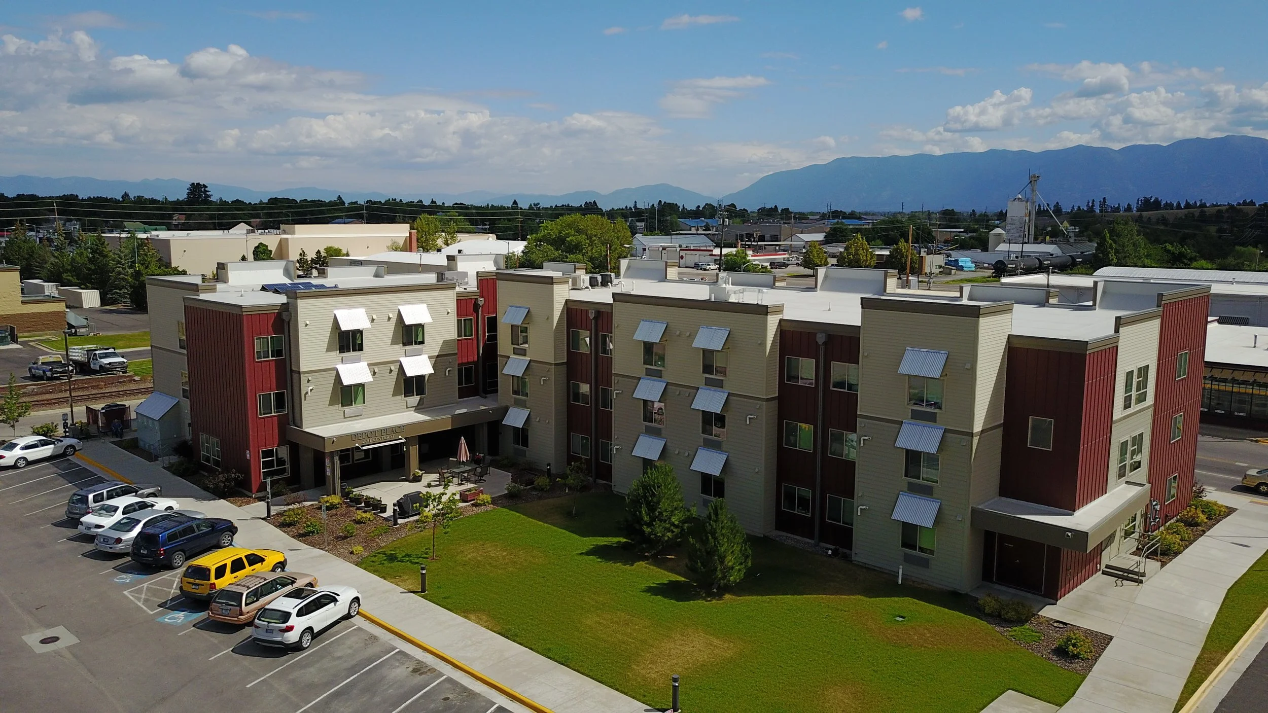 Multi-story apartment complex with green lawn and parking lot in foreground, mountains in background, under a partly cloudy sky.